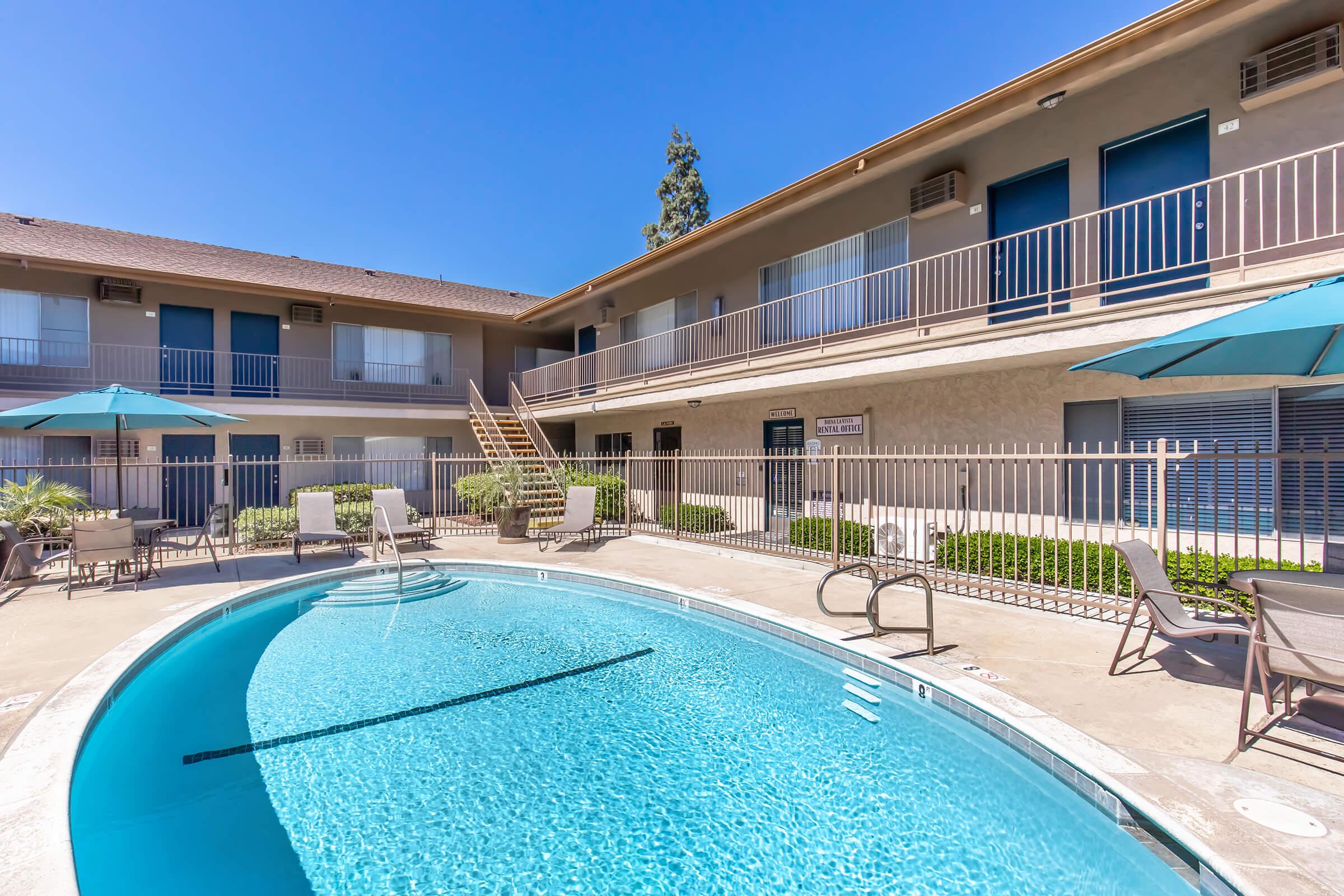 A clear blue swimming pool surrounded by a sunlit courtyard with lounge chairs and patio umbrellas. In the background, a two-story building features balcony rooms and green landscaping, creating a relaxing outdoor space.
