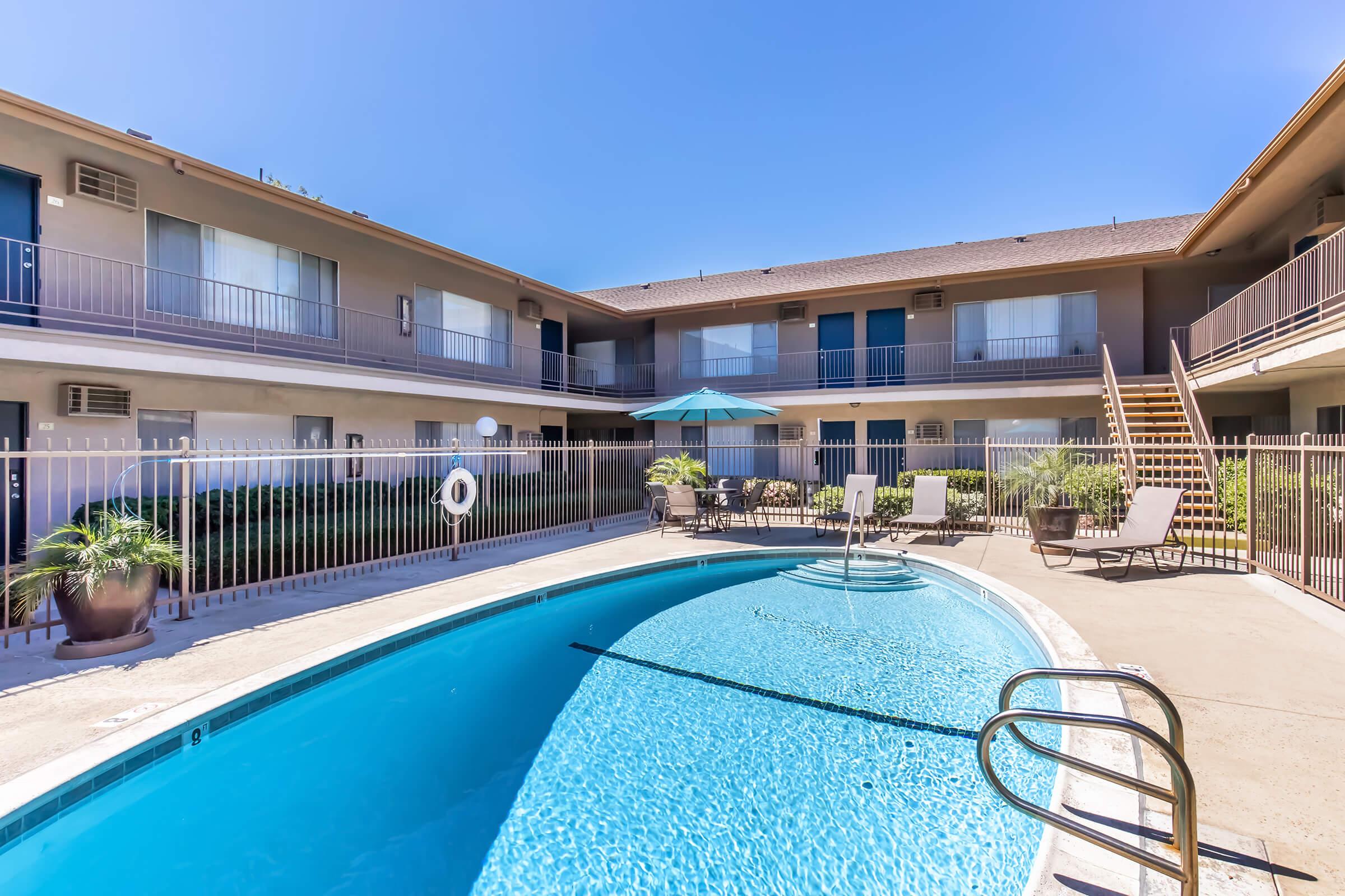A clear blue swimming pool surrounded by a well-maintained courtyard in an apartment complex. Comfortable lounge chairs and an umbrella offer a relaxing space, with two floors of balconies visible around the pool area under a bright blue sky.