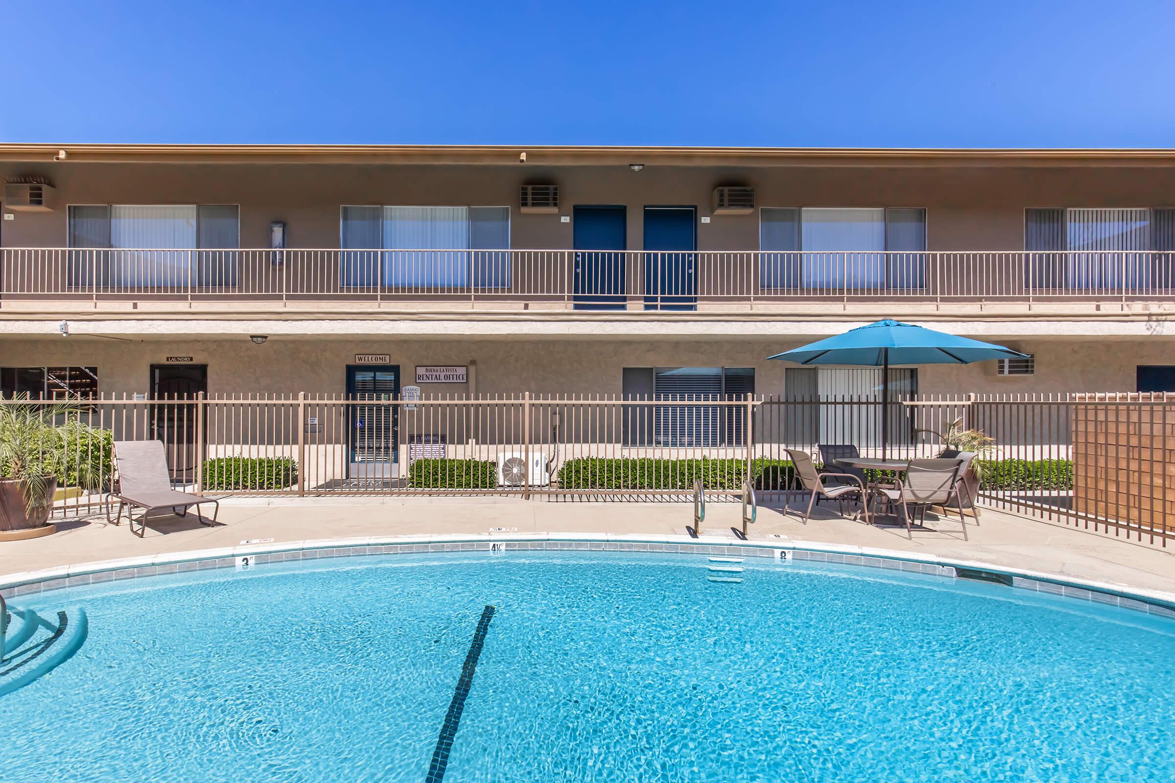 A clear view of a swimming pool in the foreground, with lounge chairs and an umbrella. In the background, a two-story building with balconies and multiple doors is visible under a bright blue sky. The area is well-maintained, with greenery around the poolside.