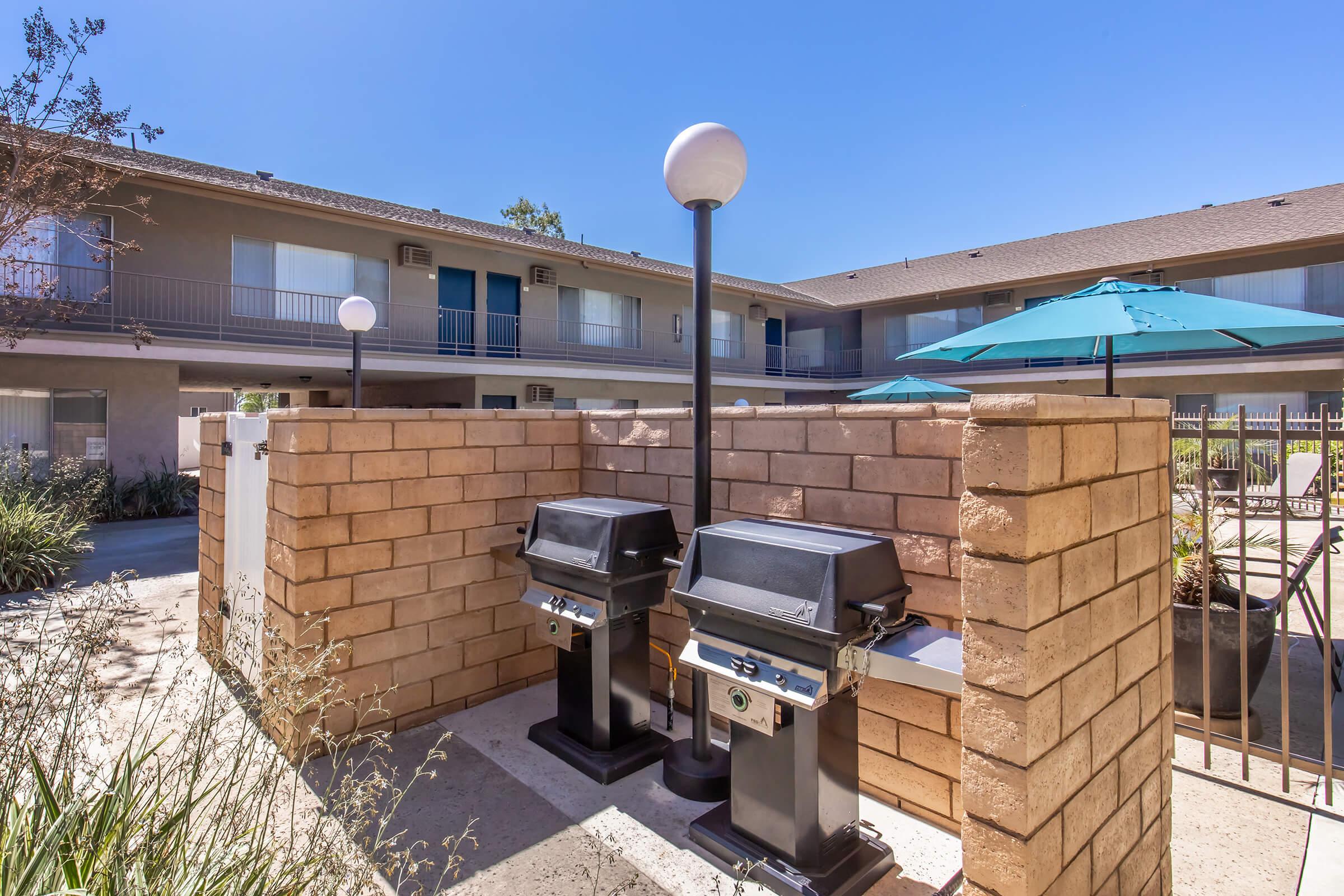 Two black barbecue grills placed between stone walls in a courtyard area. Light gray buildings with balconies are visible in the background, along with blue umbrellas providing shade. Bright blue sky adds to the outdoor ambiance.