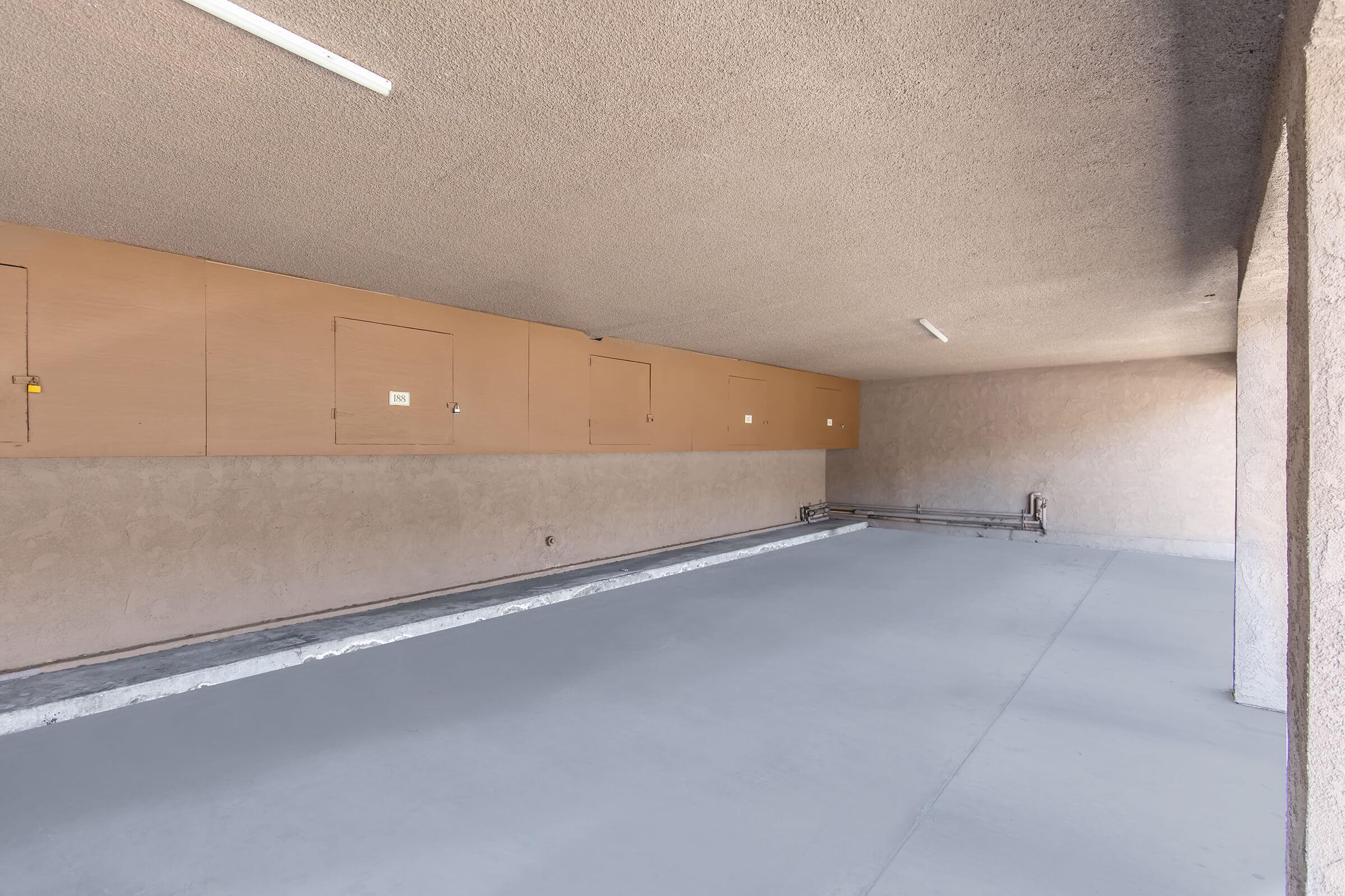 A spacious indoor area with smooth gray flooring. The walls are a light beige color, and there are several wooden storage cabinets mounted at the top along one side. The ceiling features plain light fixtures, and there is a pipe running along the floor. The overall feel is clean and minimalistic.