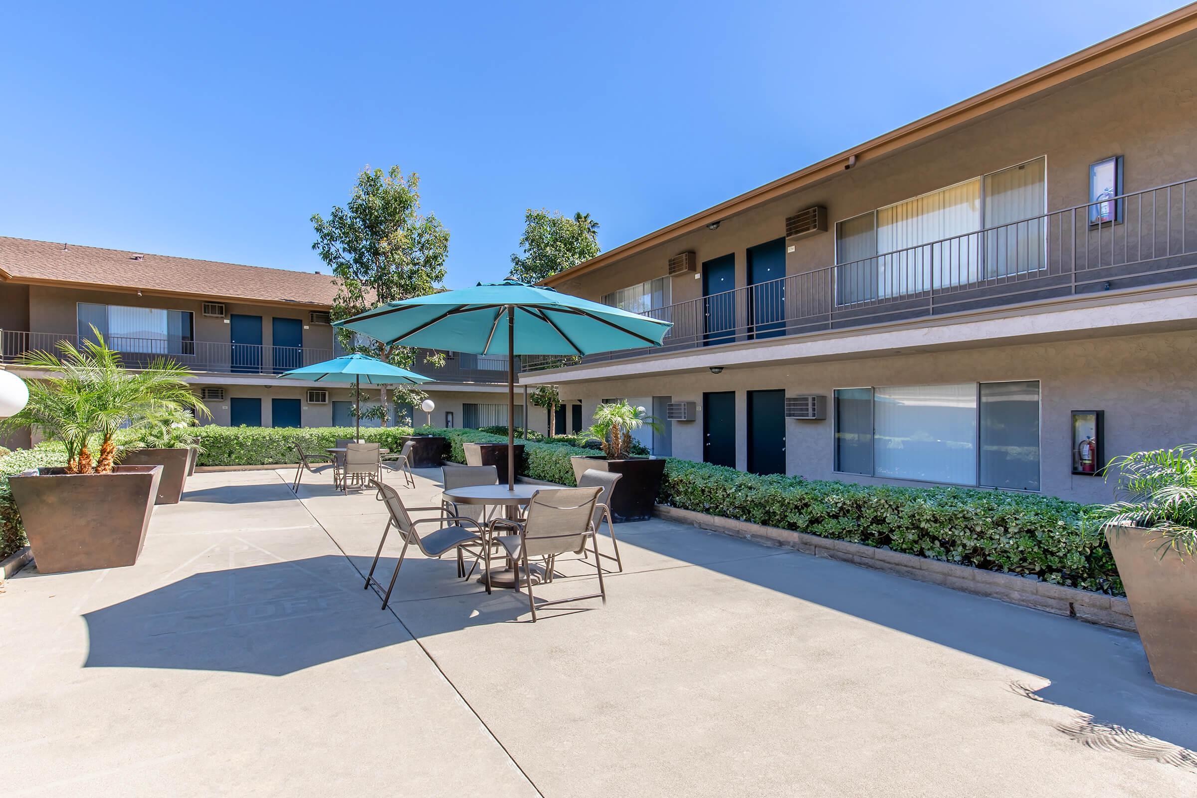 A courtyard area featuring tables and umbrellas, surrounded by potted plants and palm trees. Two-story buildings with balconies are visible in the background, under a clear blue sky. The setting suggests a relaxed outdoor space, likely for residents or guests to enjoy.