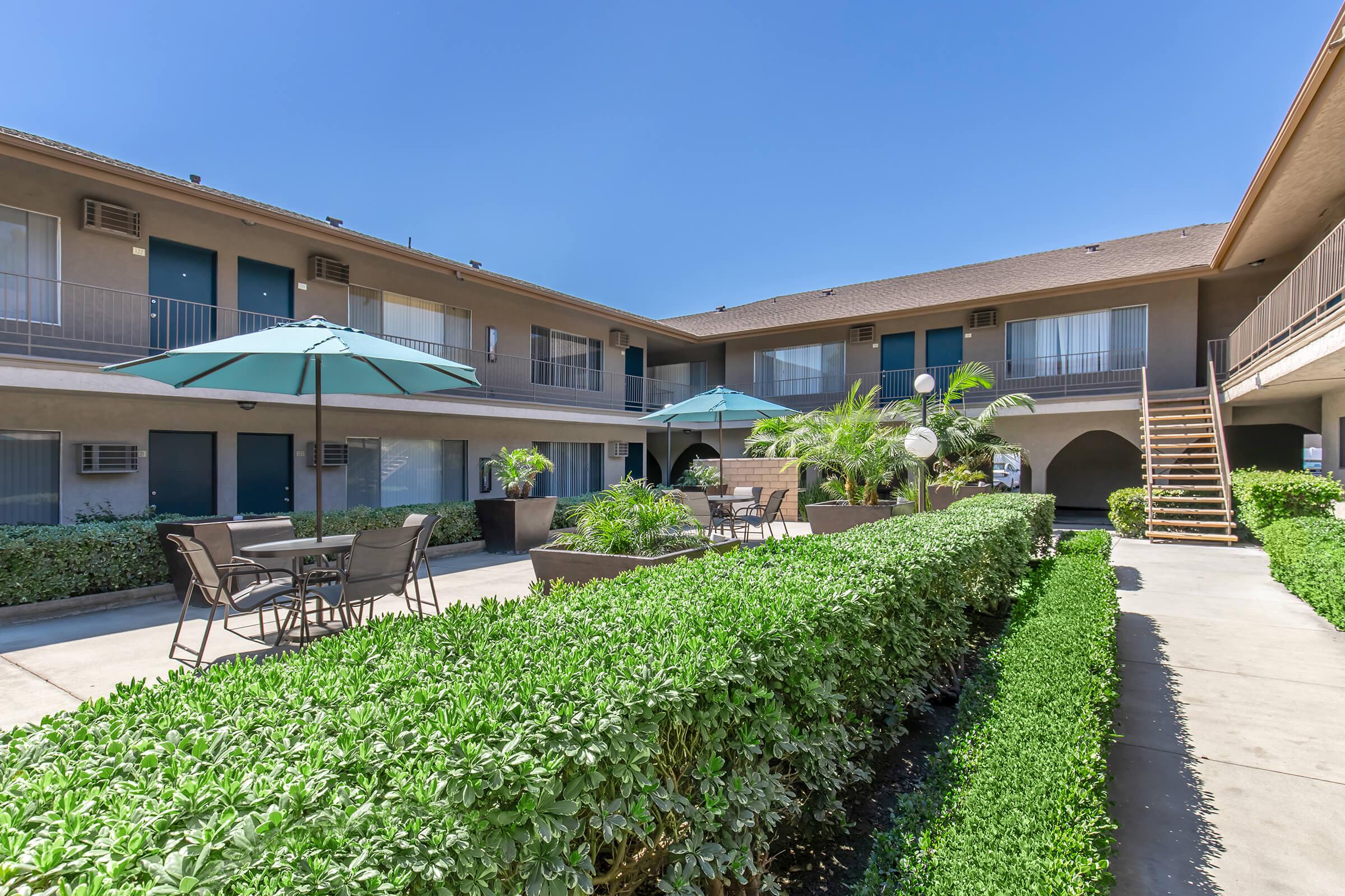A view of a courtyard area in an apartment complex, featuring lush green hedges, outdoor seating with umbrellas, and a staircase leading to upper-level units, all set against a clear blue sky. The layout suggests a welcoming outdoor space for residents to relax.