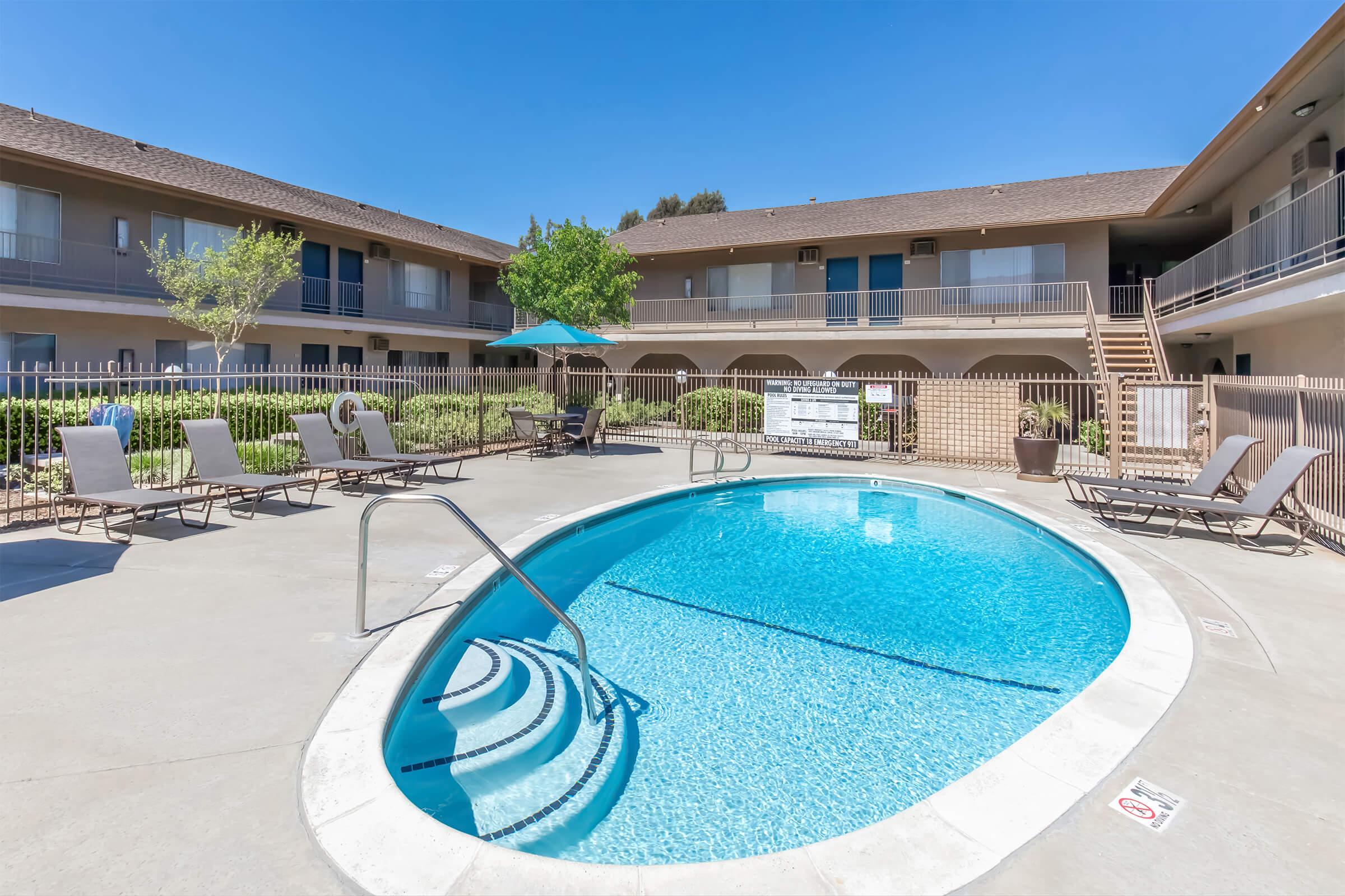 A clear swimming pool surrounded by lounge chairs, with a shaded area under an umbrella. The pool is located in a courtyard of a two-story building, with landscaped greenery and steps leading to the upper level in the background. The sky is bright and blue, indicating a sunny day.