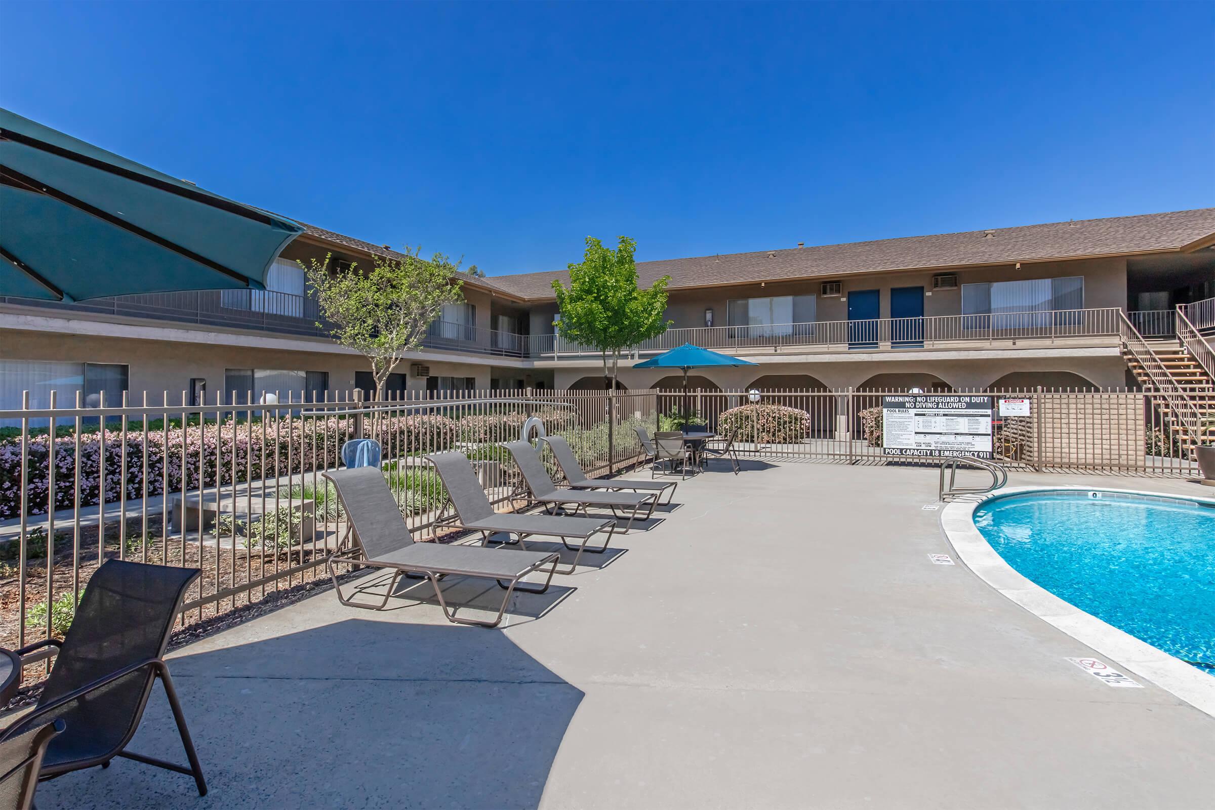 A pool area featuring lounge chairs and umbrellas, surrounded by a gated fence. In the background, a two-story building with balconies is visible under a clear blue sky. The setting creates a relaxed atmosphere ideal for leisure and enjoyment.