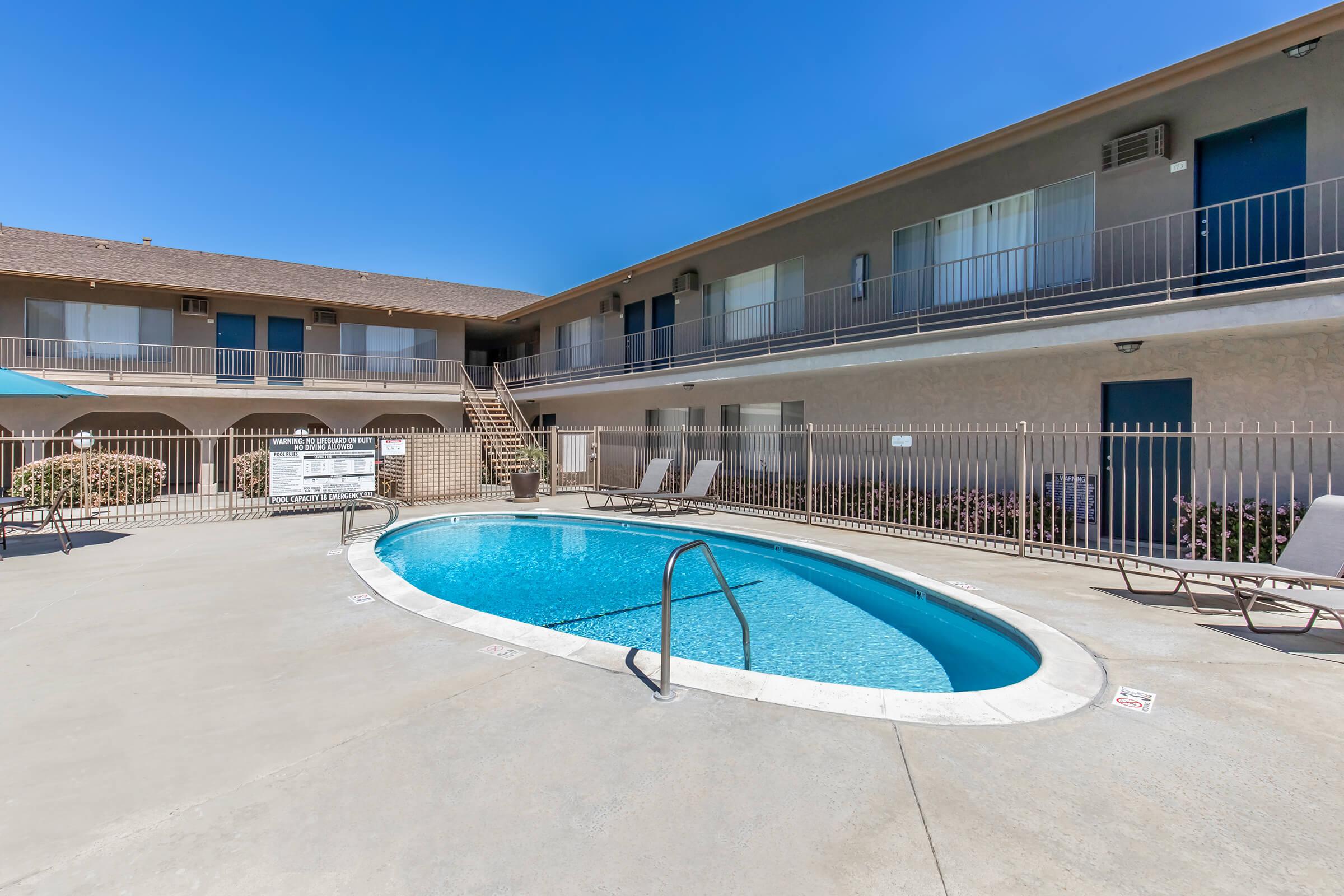 A clear blue swimming pool surrounded by lounge chairs, located in a courtyard of a two-story building with multiple balconies and doors. The sky is bright blue, and the area is well-lit, creating a welcoming atmosphere for relaxation.