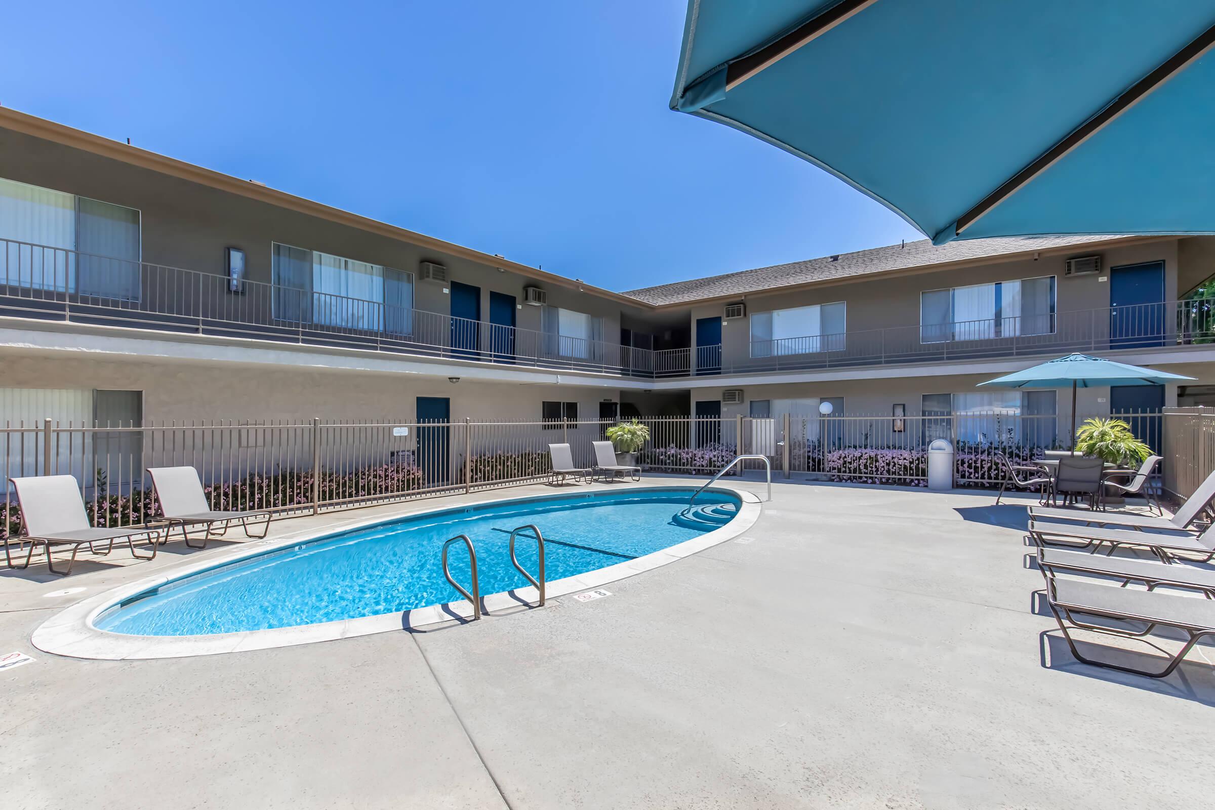 A clear blue swimming pool surrounded by a deck with lounge chairs, set against a backdrop of a two-story building with balconies. An umbrella provides shade, and the sky is bright and sunny. Lush greenery adds a touch of color to the scene, creating a relaxing poolside atmosphere.