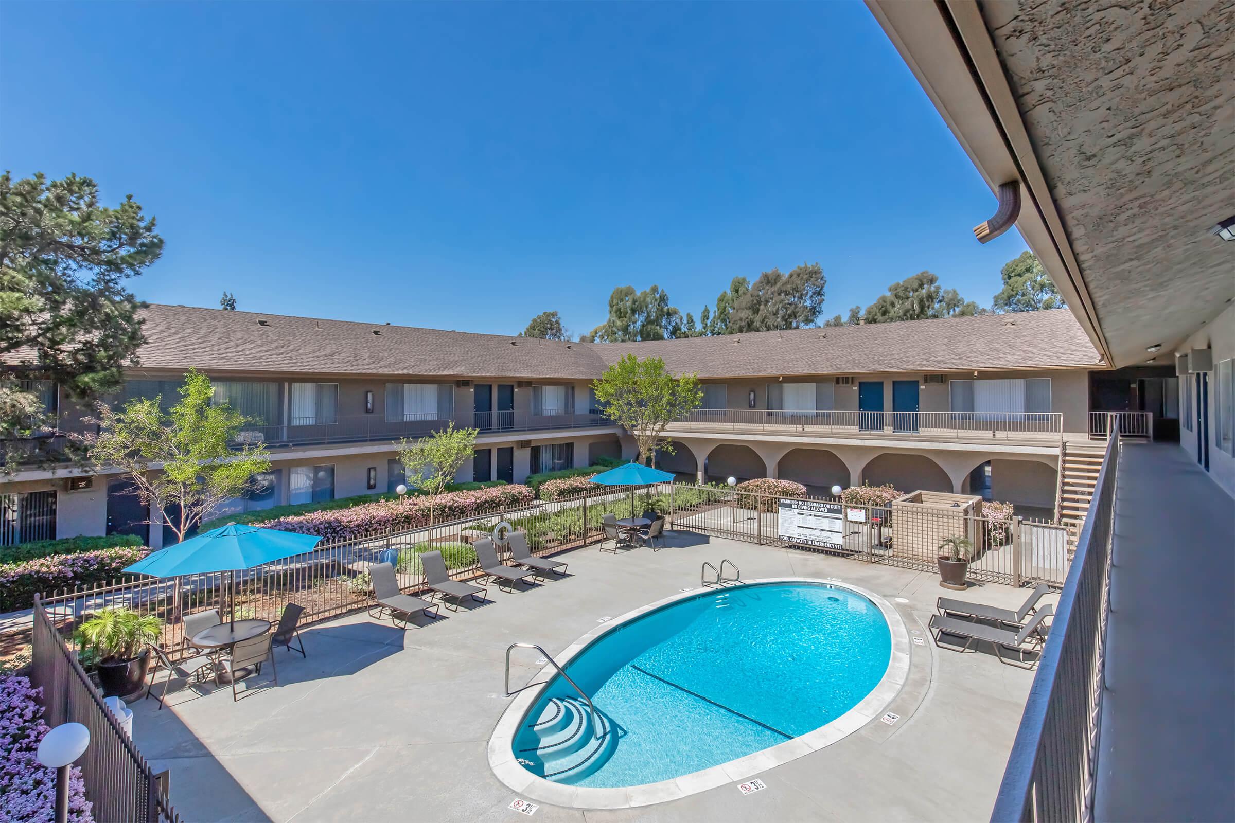 A sunny courtyard featuring a clear blue swimming pool surrounded by lounge chairs and umbrellas. The pool area is bordered by colorful flowerbeds and two-story buildings with balconies overlooking the pool. Lush trees are present, and the sky is bright blue with no clouds.