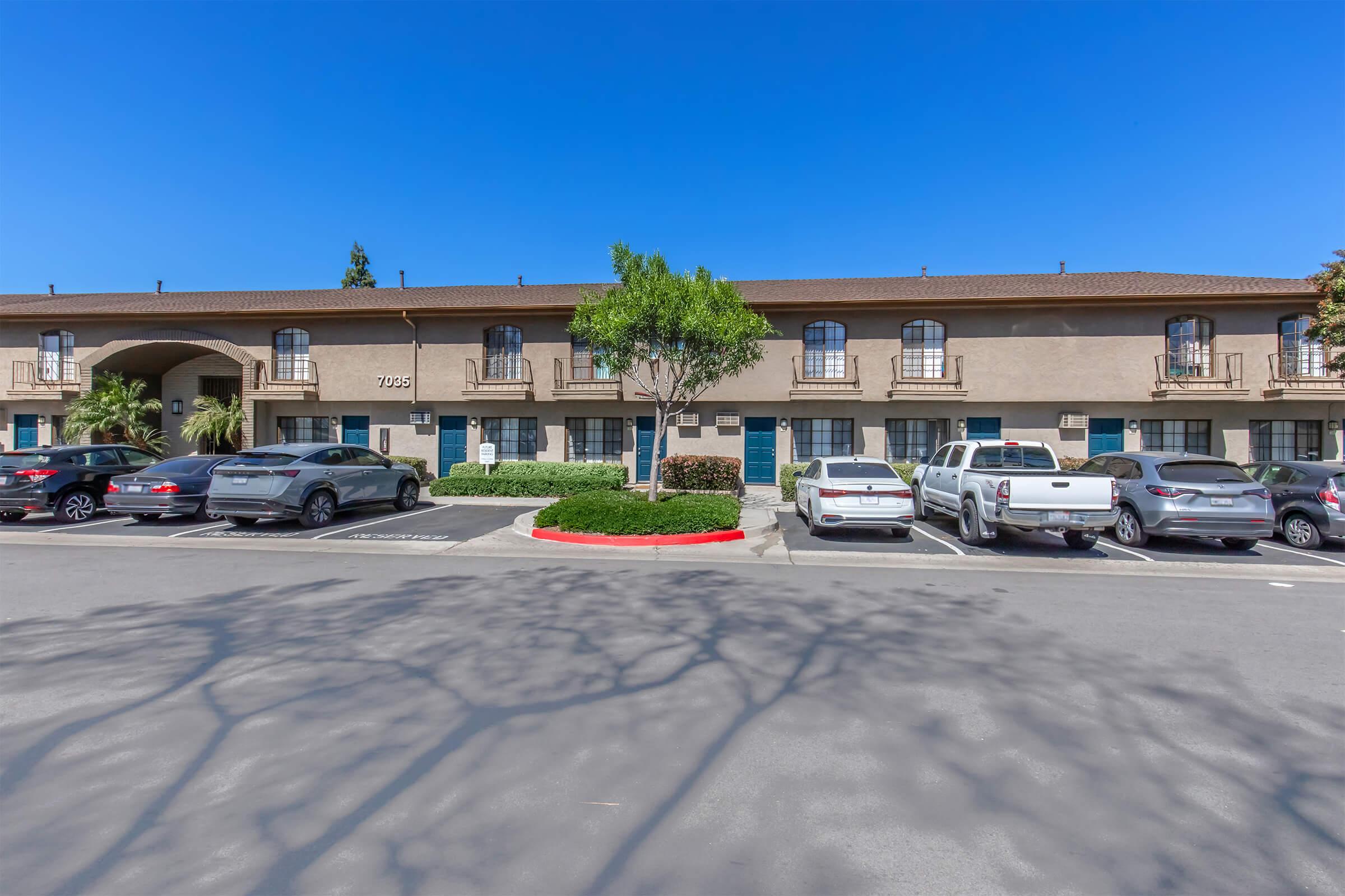 A multi-unit building with two stories, featuring several balconies and multiple windows. In the foreground, there is a parking lot with several vehicles parked, including sedans and trucks. The sky is clear and blue, and there are lush green plants surrounding the area.
