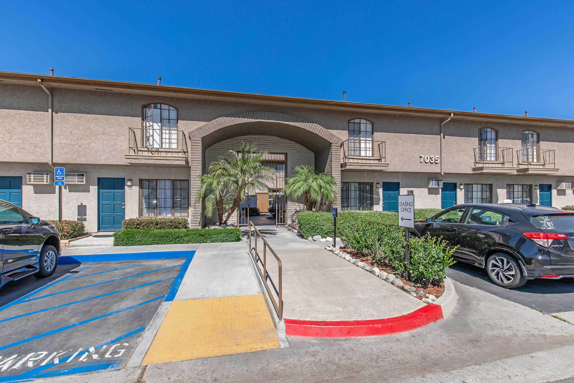 Exterior view of a two-story apartment building featuring a central entrance with palm trees on either side, a blue sky above, and parking spaces in the foreground. The pathway includes a wheelchair-accessible ramp leading to the entrance.