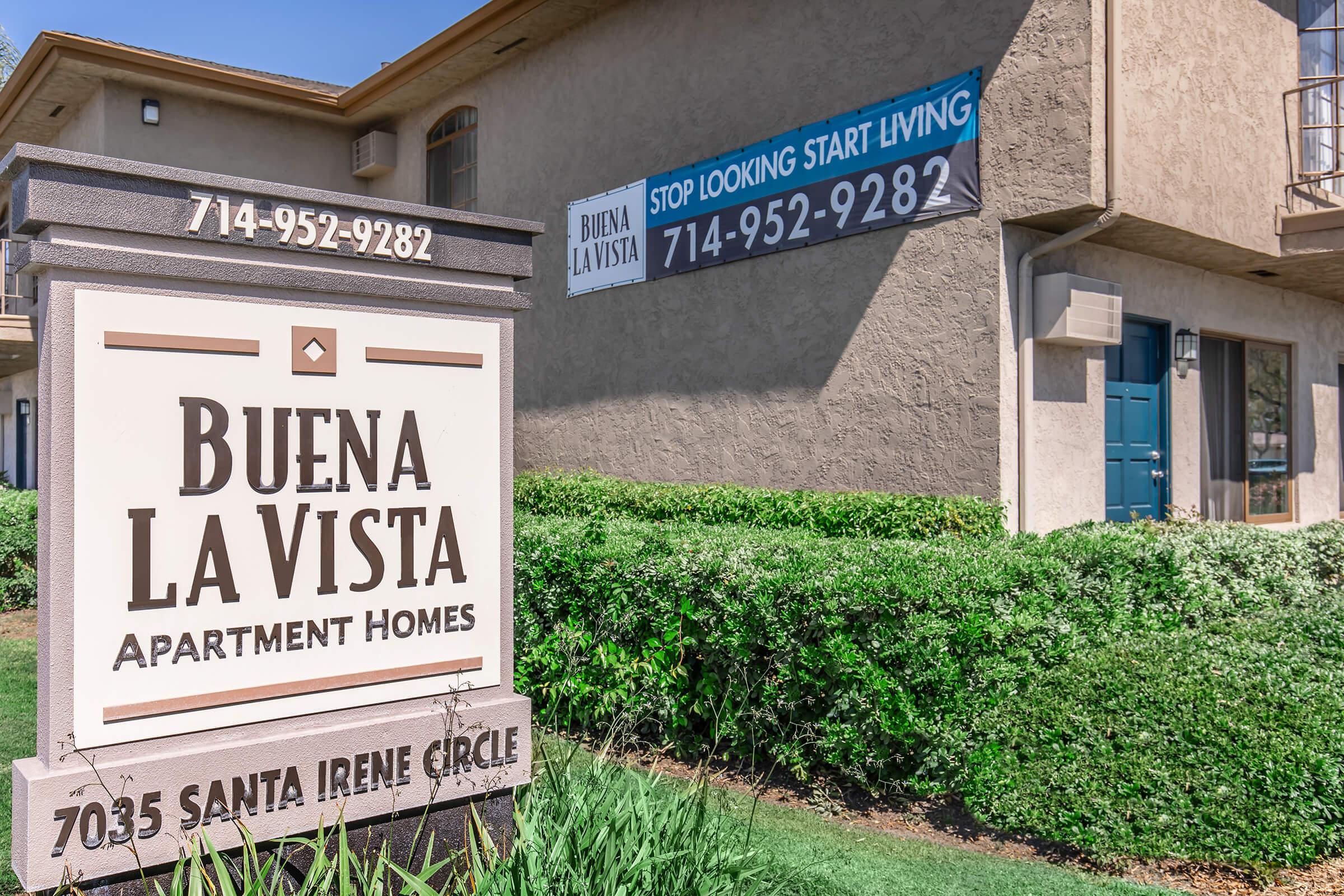Sign for Buena La Vista Apartment Homes with contact information, located at 7035 Santa Irene Circle. The sign includes a phone number (714-952-9282) and an advertisement encouraging potential residents to stop looking and start living. The building features a beige exterior and landscaping.