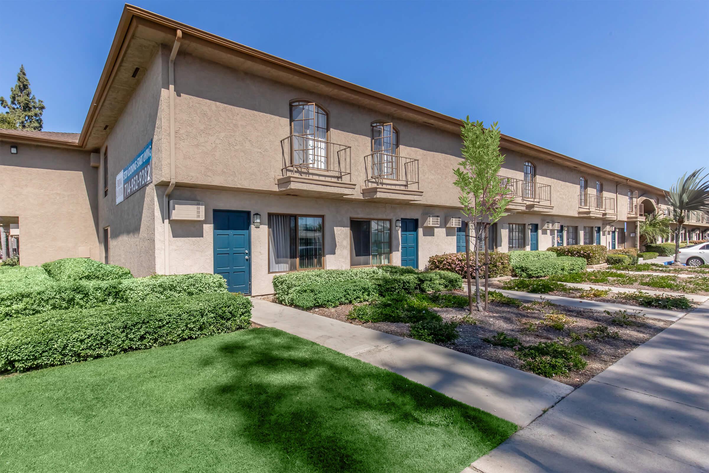 A view of a two-story building with a light brown exterior. The facade features several windows with decorative balconies. The well-maintained landscaping includes green shrubs and grass. Clear blue skies are visible above.