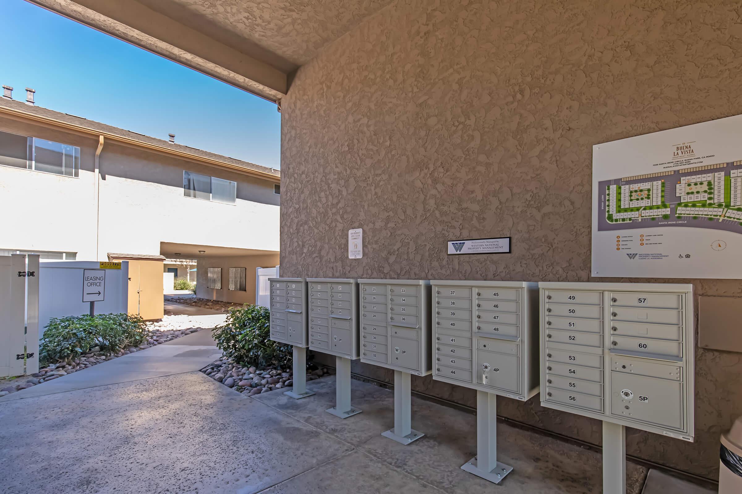A corridor of an apartment complex featuring a row of individual mailboxes on the left. On the right, a map or directory of the complex is displayed. In the background, a pathway leads to other buildings with shrubs and a clear blue sky above.