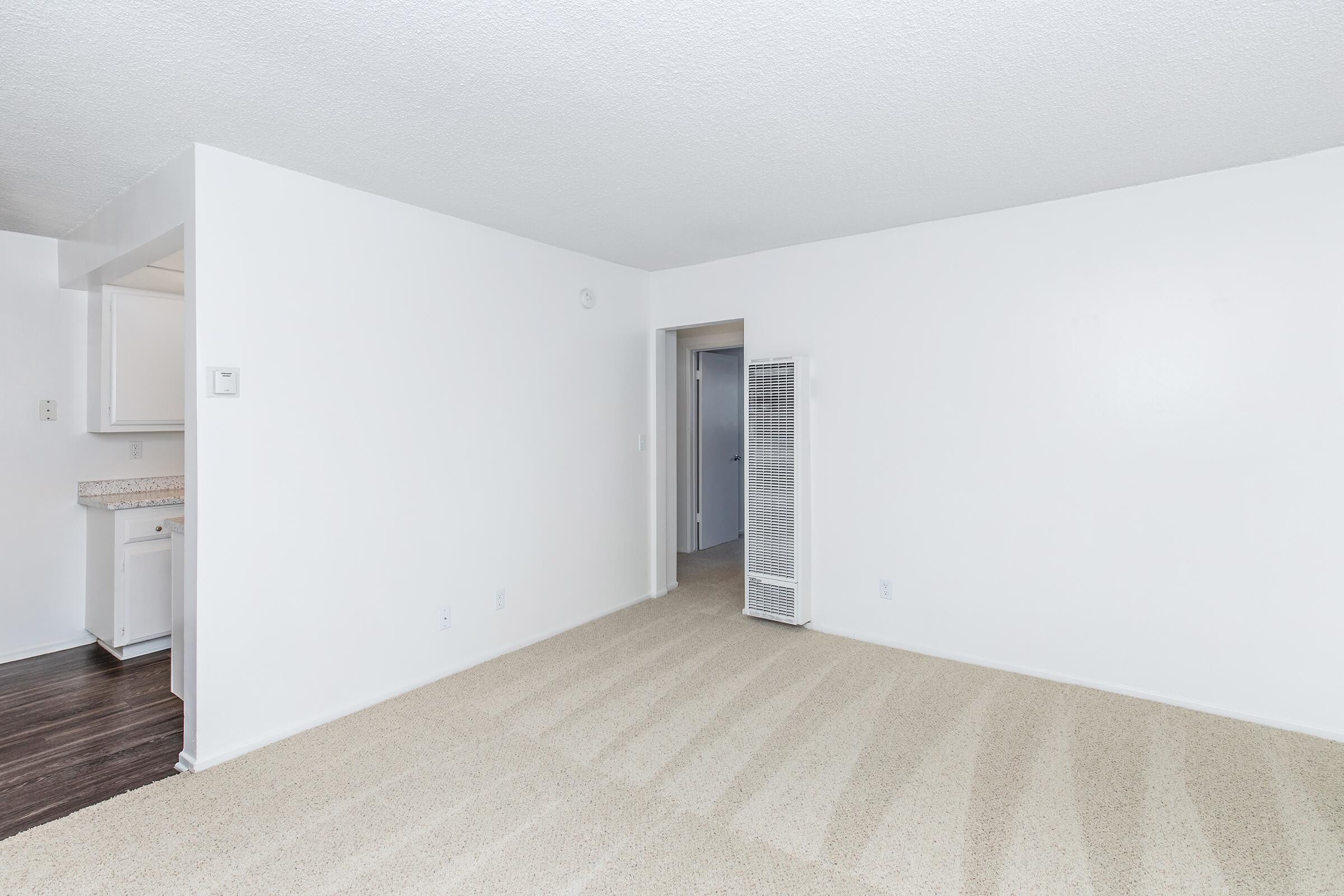 A spacious, well-lit living area with beige carpet flooring and white walls. To the left, there's a kitchen area with a countertop. A door leads to another room, and a freestanding white fan is positioned against the wall. The overall atmosphere is clean and inviting.