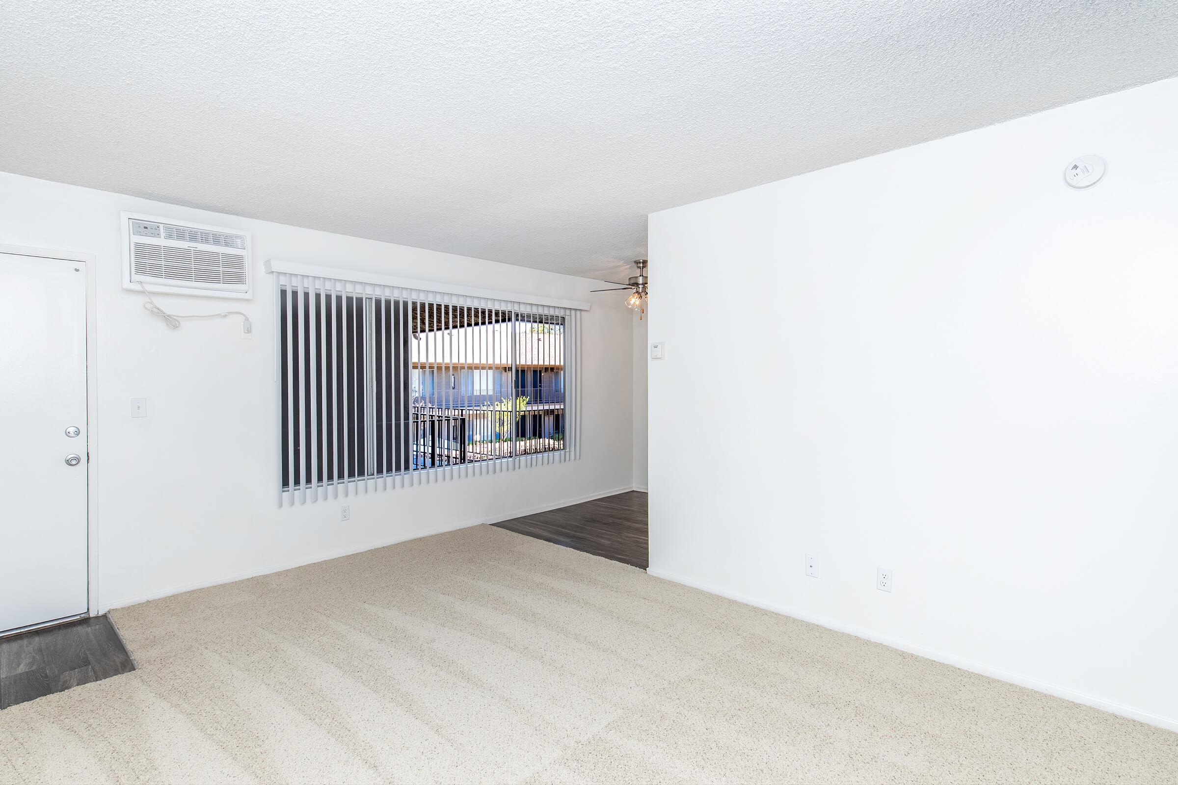 Bright, empty living room featuring light beige carpet, white walls, and vertical blinds covering a large window. Air conditioning unit on the wall and a ceiling fan visible. The space is tidy and open, creating a welcoming atmosphere.