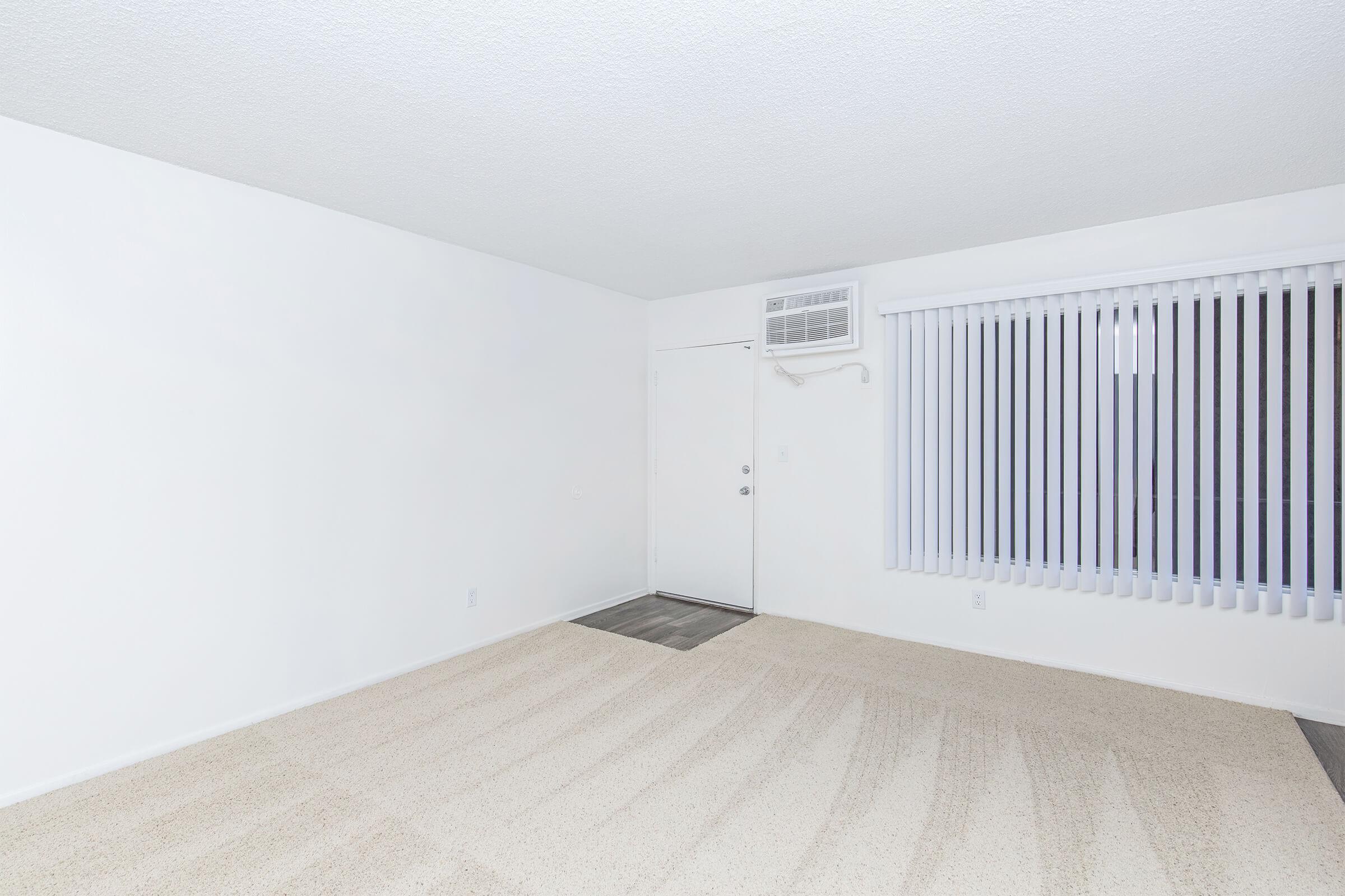 A clean, empty room with white walls and a textured ceiling. The floor is covered in light beige carpet with subtle stripes. There's an air conditioning unit mounted above a window with vertical blinds, and a door leading outside is visible on the left side of the image. The overall ambiance is bright and minimalist.