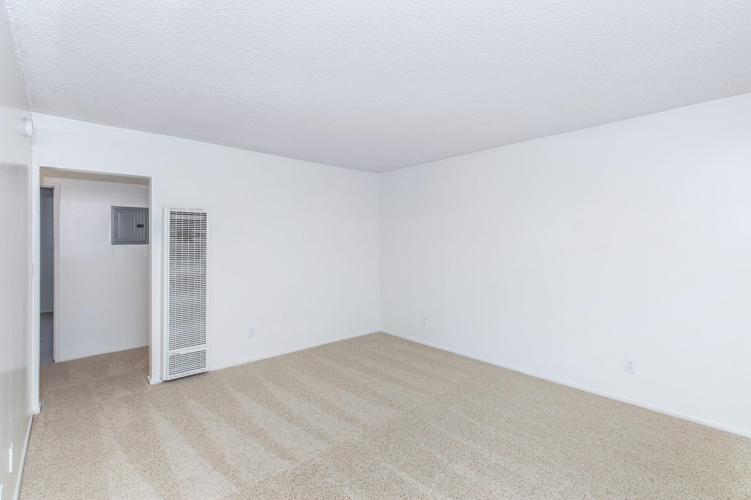 Empty room with beige carpet and white walls, featuring a doorway on the left leading to another room. An air vent is visible on the right wall. The space is bright and uncluttered, suggesting it is a living area or apartment interior.
