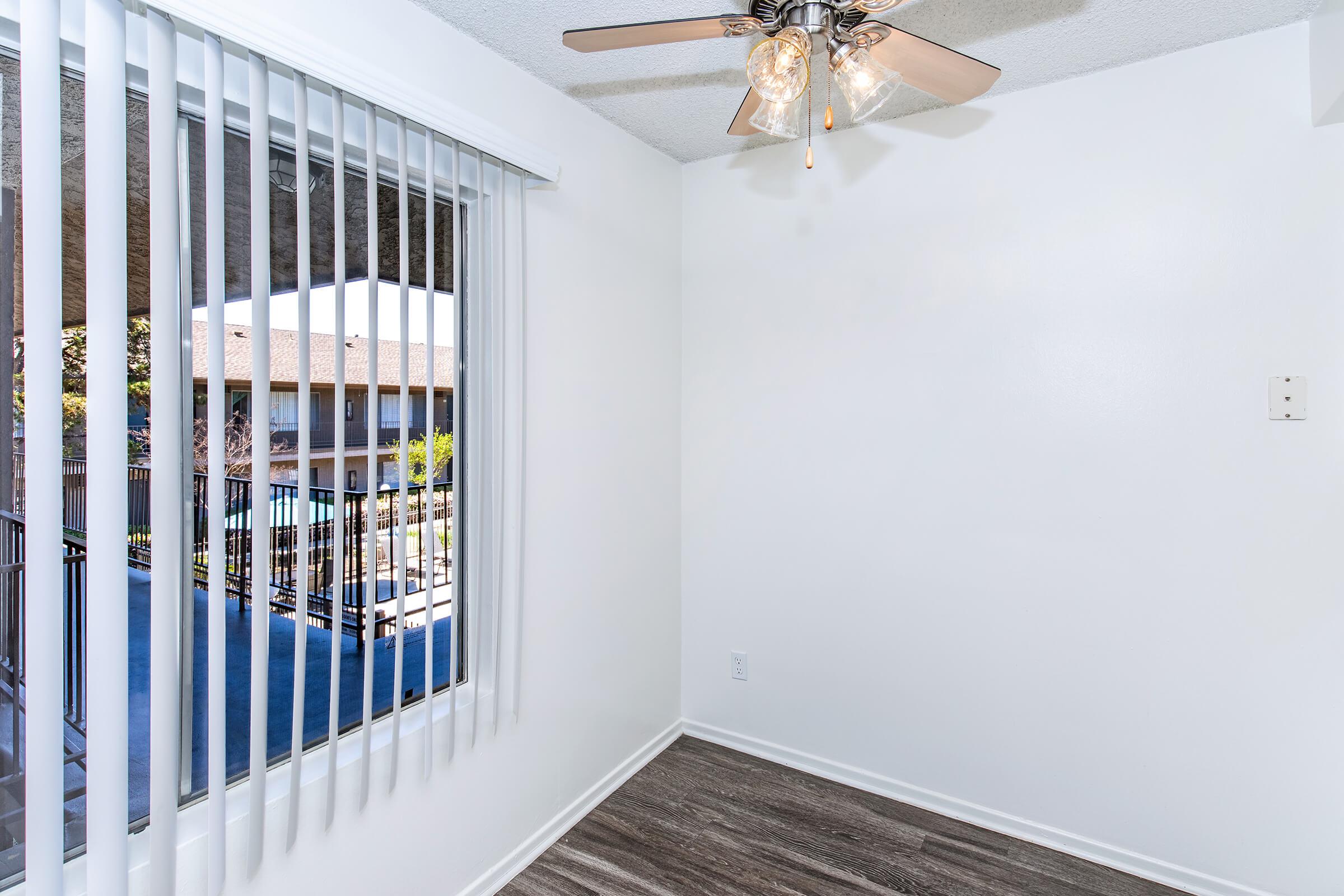 A small, bright room with a ceiling fan. There are vertical blinds covering a large window that overlooks a balcony area. The floor features wood-like laminate, and the walls are painted white, creating a clean and modern appearance.