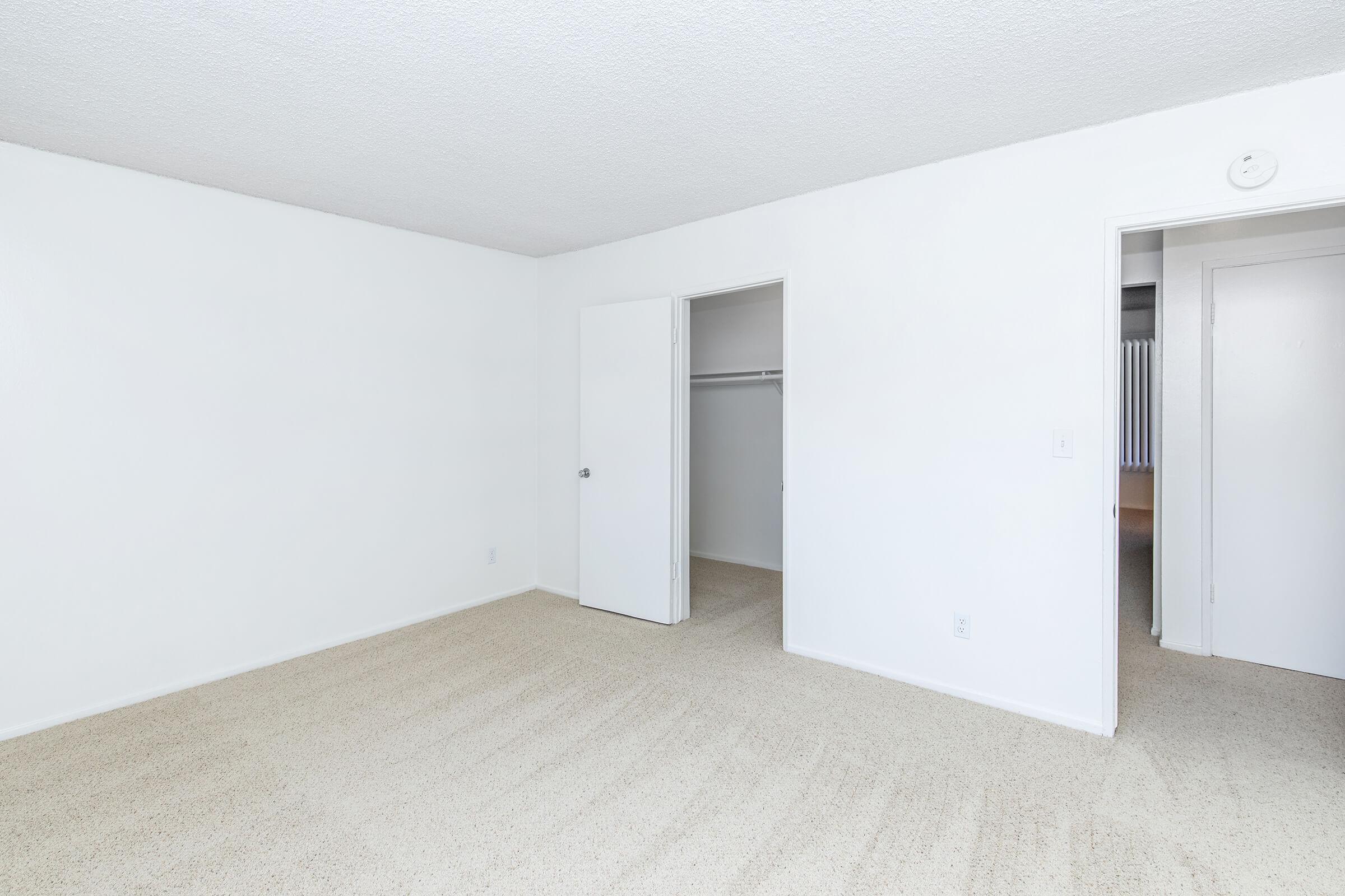 A simple, empty room with light-colored walls and carpet. There are two doors: one leading to a closet and the other to another space. Natural light brightens the room, highlighting its minimalist design. The space is clean and versatile for various uses.