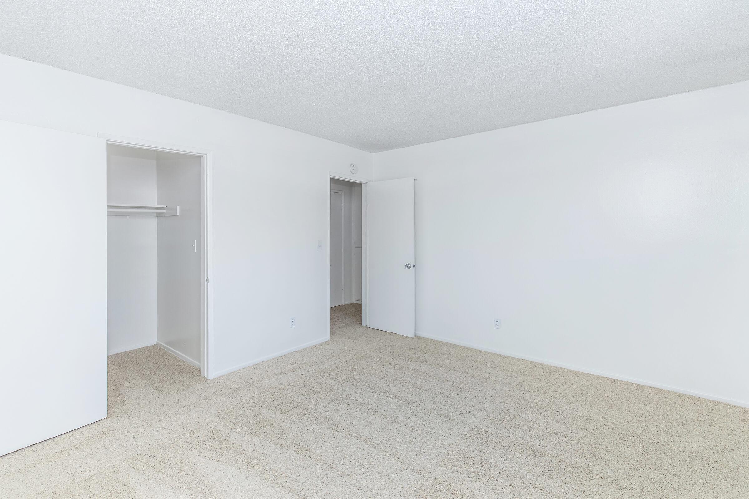 A bright and spacious empty room with light-colored walls and a textured ceiling. It features a doorway leading to a small closet on the left, with another door on the right. The floor is covered with beige carpeting, creating a clean and minimalist atmosphere.
