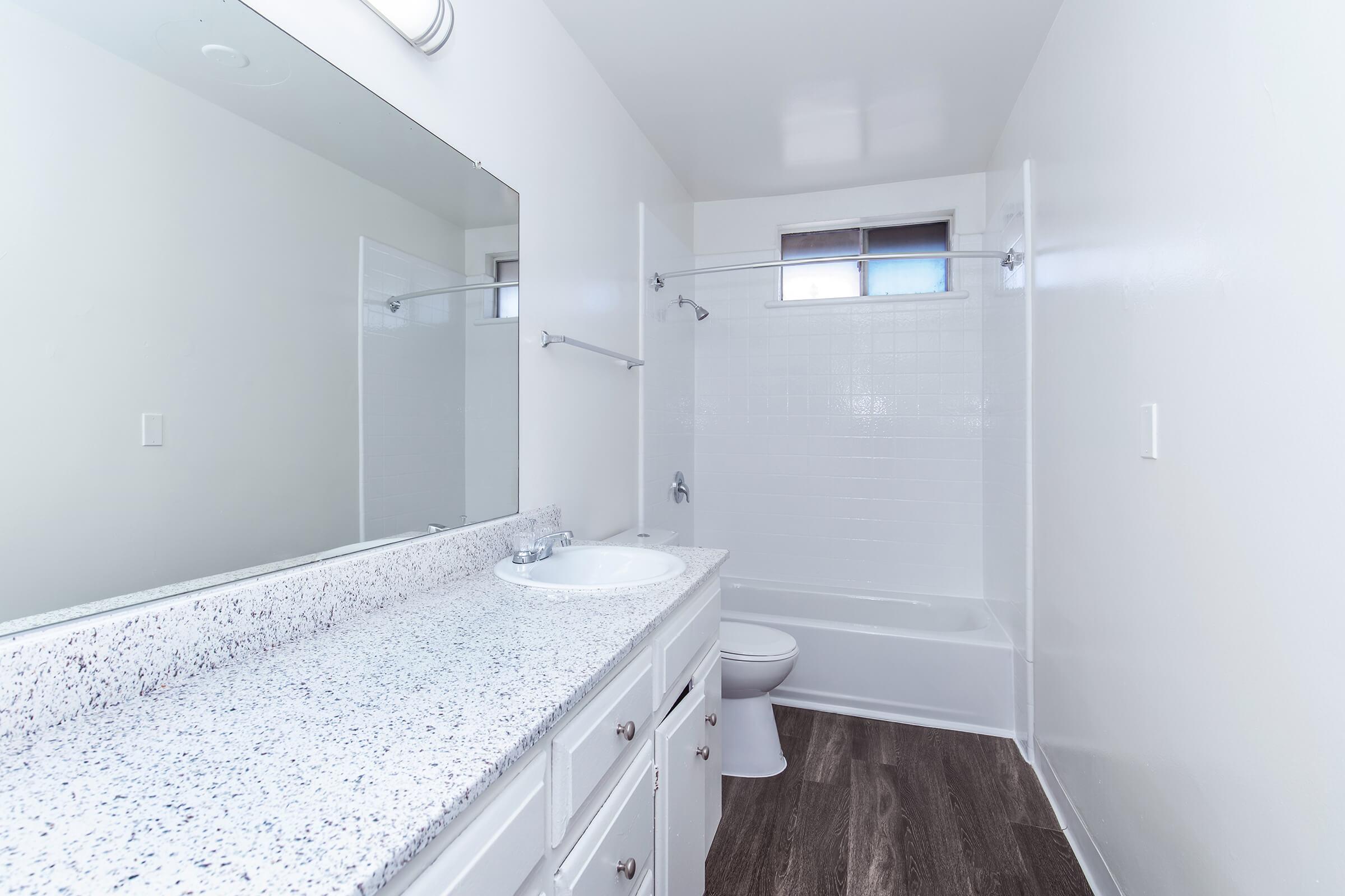 A clean, modern bathroom featuring a white bathtub with shower, a single sink with a speckled countertop, and a large mirror above the sink. Light-colored walls and a dark wood-like floor create a bright and spacious atmosphere. A window allows natural light to enter the room.