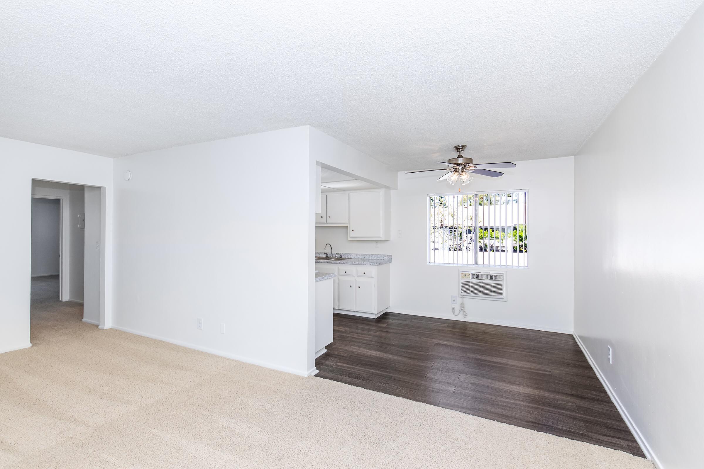 Bright and spacious living area with a combination of carpet and hardwood flooring. Features include a ceiling fan, a window with blinds, and an open view into a kitchen area with white cabinetry. The walls are painted in a light color, enhancing the airy feel of the room.