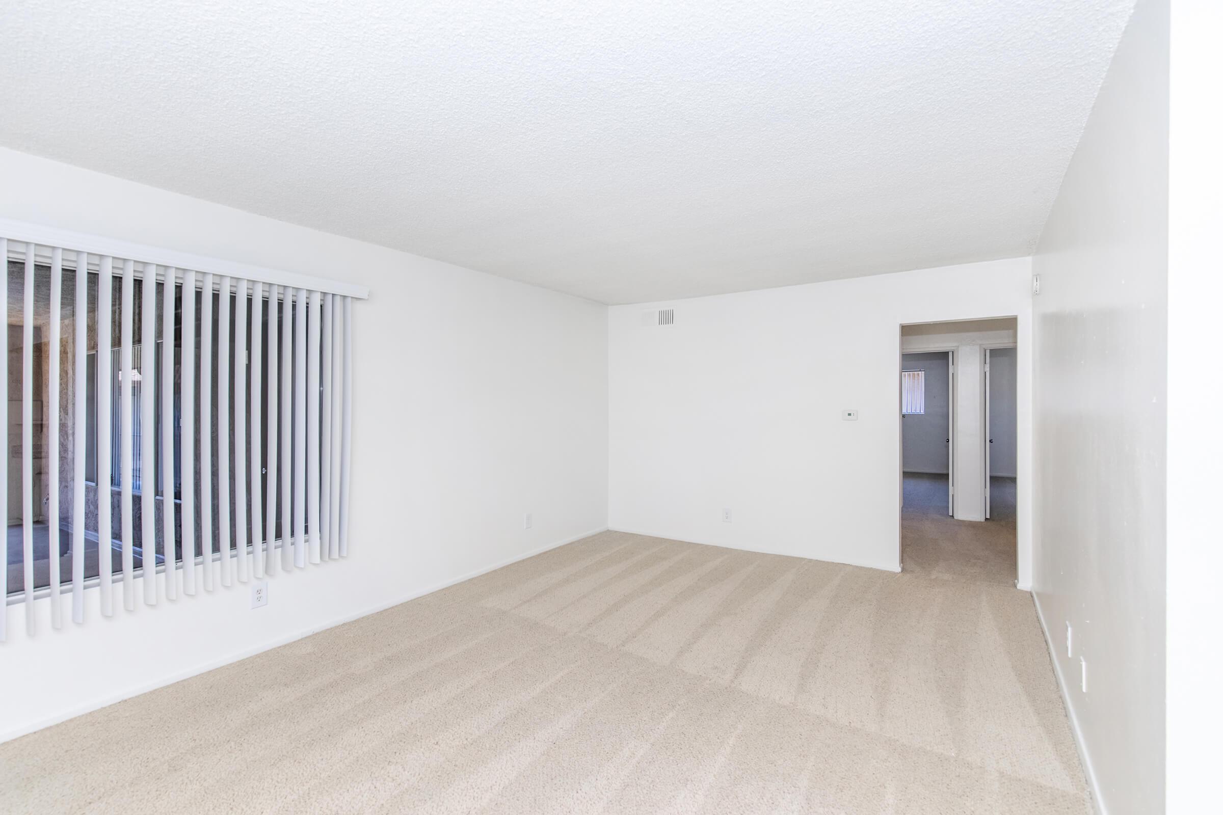 An empty living room with beige carpet and white walls. There's a large window with vertical blinds allowing natural light, and a doorway leading to another room. The space appears clean and well-maintained, ideal for personalizing with furniture and decor.