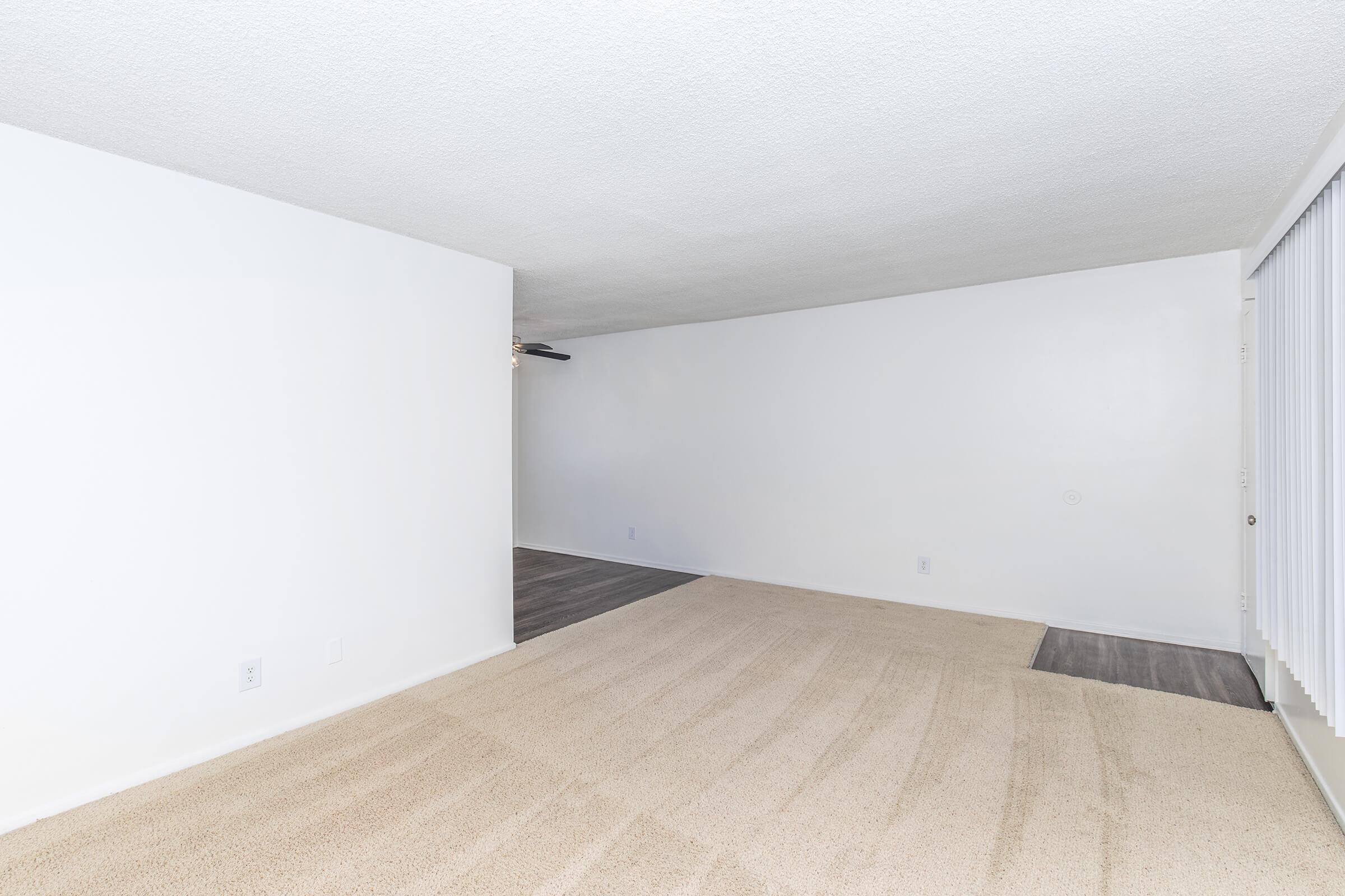 A spacious, empty living room with beige carpet and white walls. There is a ceiling fan on the left side and a doorway leading to another room on the right. The area is well-lit, creating a bright and open atmosphere.