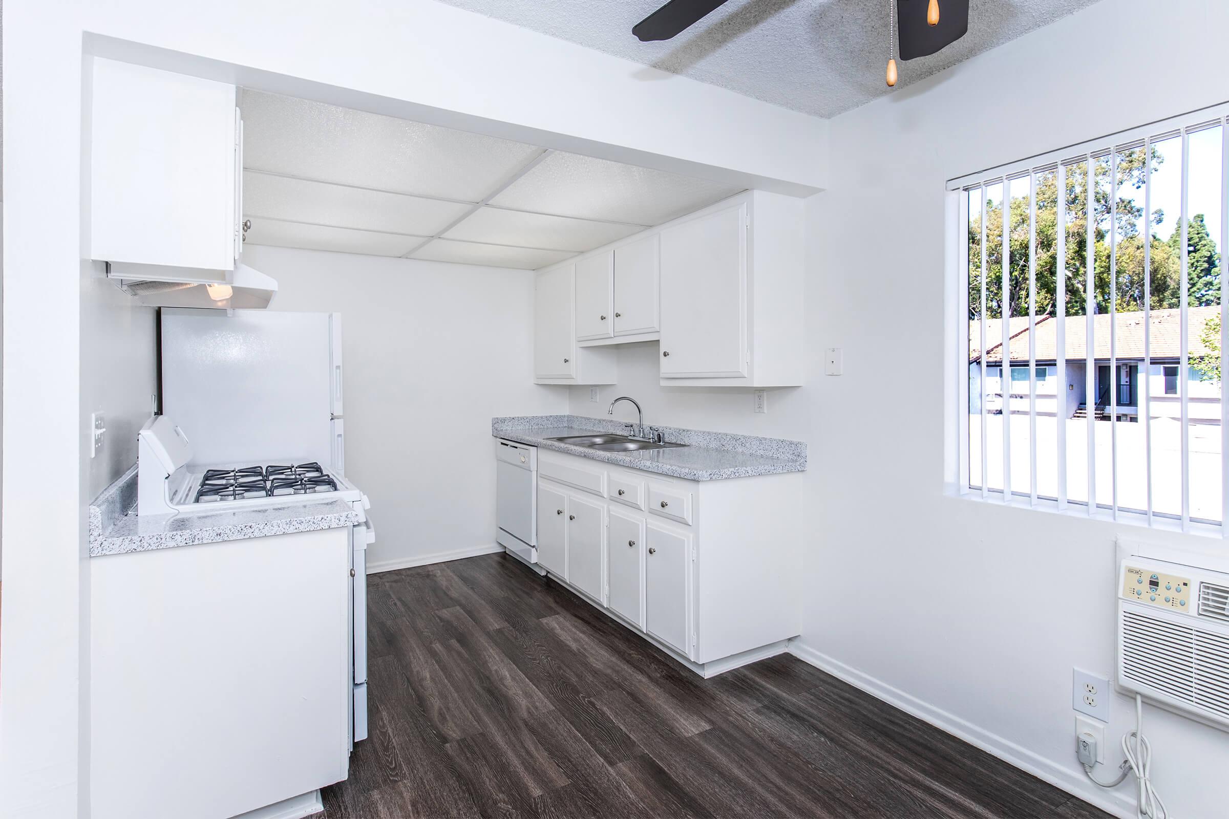 Bright, modern kitchen featuring white cabinets, a granite countertop, and a gas stove. The space includes a sink and has an open layout with natural light coming through a large window. The flooring is dark wood, adding contrast to the light-colored walls and cabinetry.