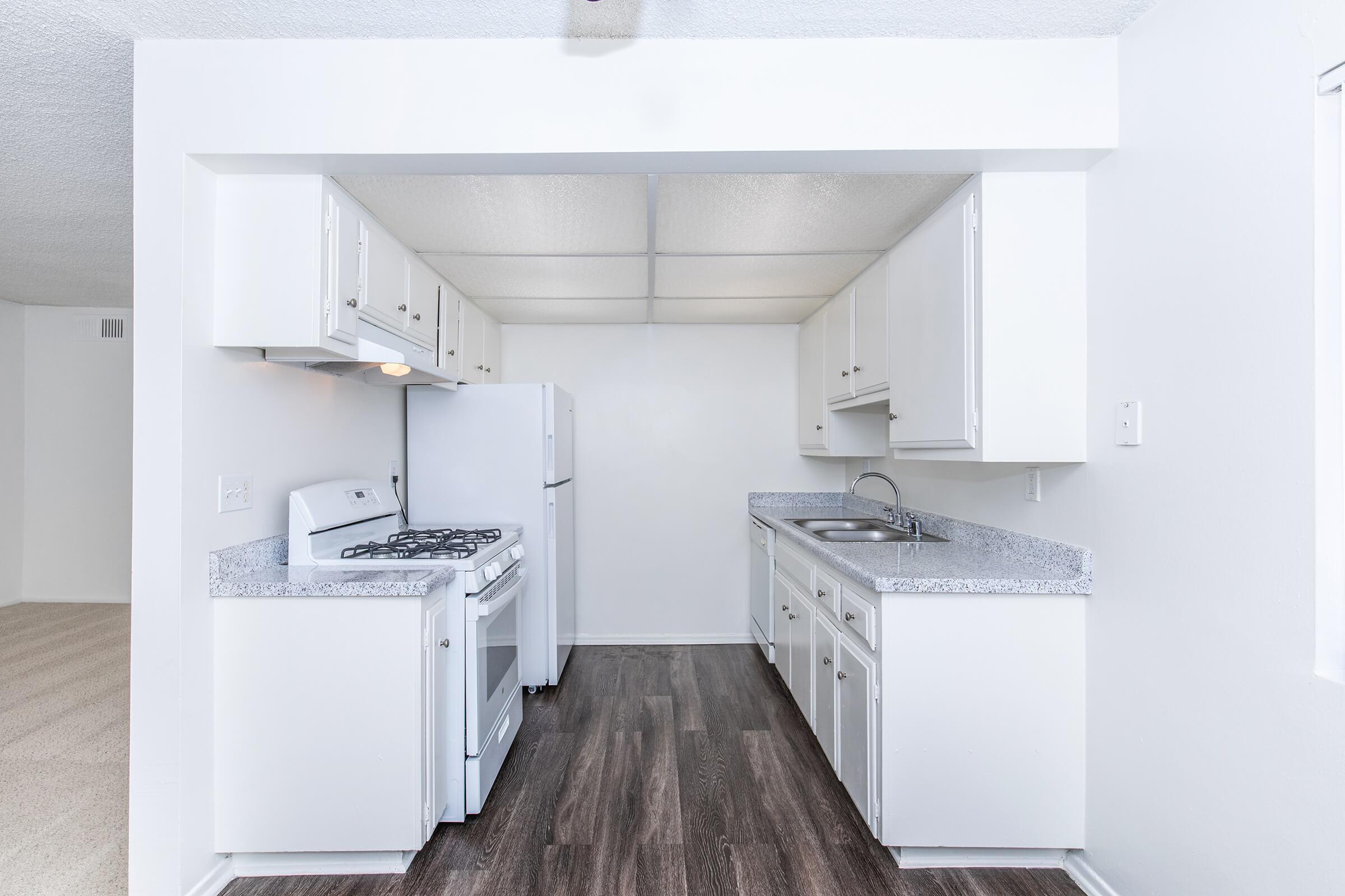 A bright, modern kitchen featuring white cabinetry, a gas stove, and a refrigerator. The countertop is gray with a sink and faucet. The room is well-lit with natural light and has a neutral-colored floor, creating a clean and spacious feel. The adjoining living area is partially visible in the background.
