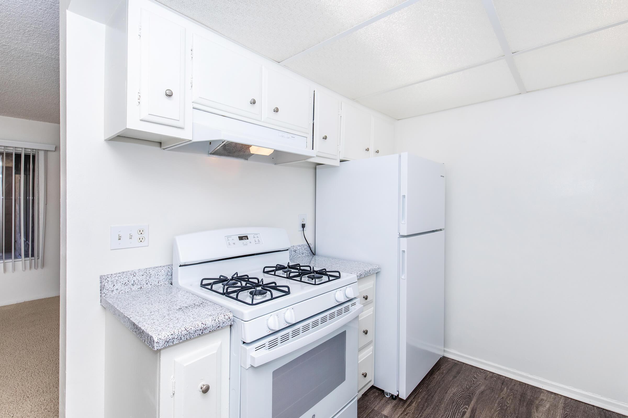 A modern kitchen featuring white cabinetry, a gas stove with an oven, and a large refrigerator. The countertops are light-colored granite, and there is a ceiling with tiles. The flooring appears to be dark laminate. The kitchen is clean and well-lit, giving it a minimalist look.
