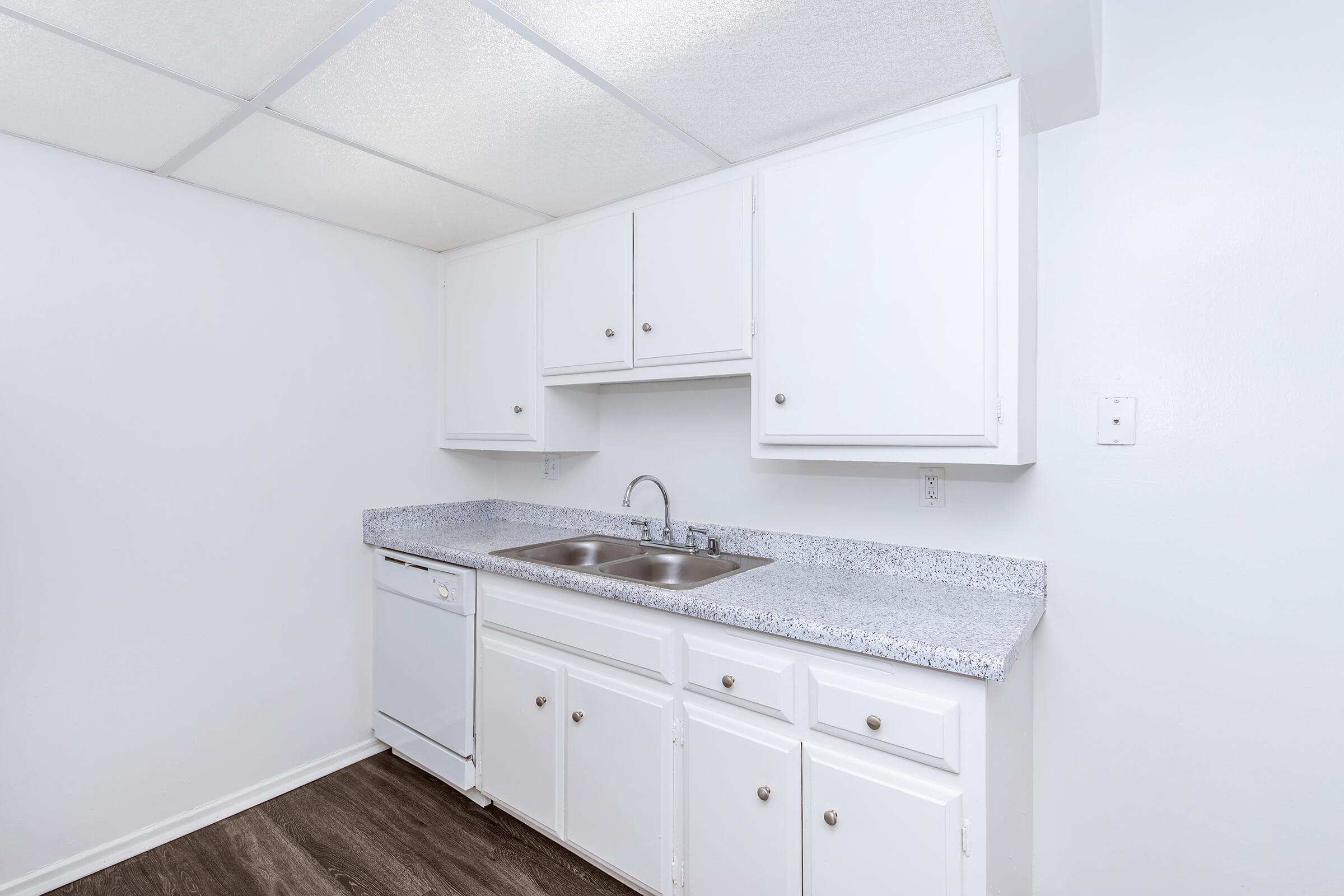 A clean, modern kitchen with white cabinets and a gray countertop, featuring a double sink and a dishwasher. The walls are painted white, and the flooring is dark laminate. The ceiling is plain and slightly textured, with no visible appliances besides the dishwasher.
