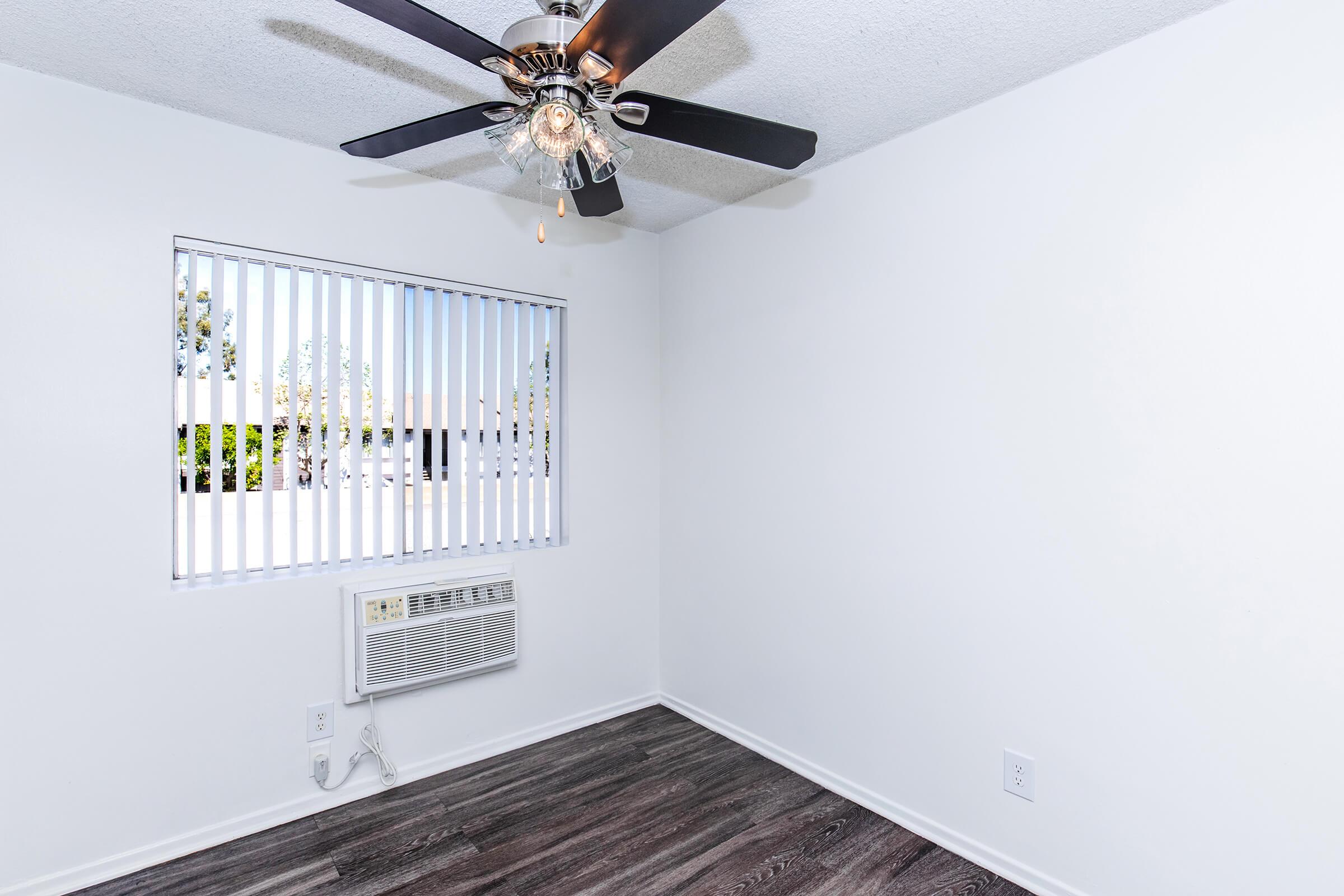 A small, empty room with white walls and a ceiling fan. There is a window with horizontal blinds letting in natural light, and an air conditioning unit mounted below the window. The floor is finished with dark wood laminate, creating a modern and clean appearance.