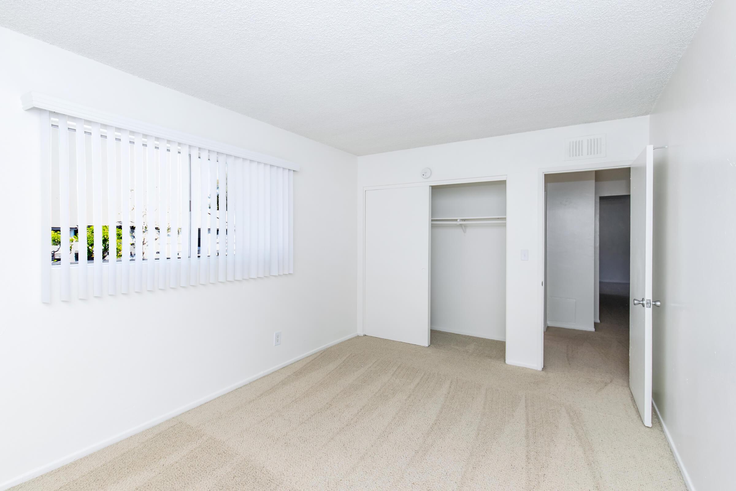 Empty room with light-colored walls and carpet. Features include a window with white blinds, a closet with sliding doors, and an open door leading to another space. The room is well-lit, giving a spacious and clean appearance.