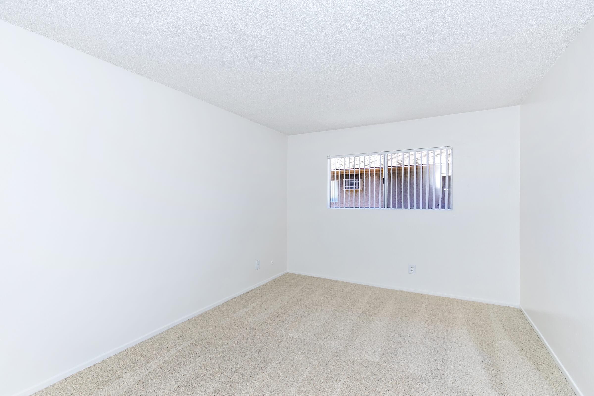 Empty room with light-colored carpet and white walls. A small window with metal bars allows natural light to enter. The space appears clean and uncluttered, suitable for various uses.