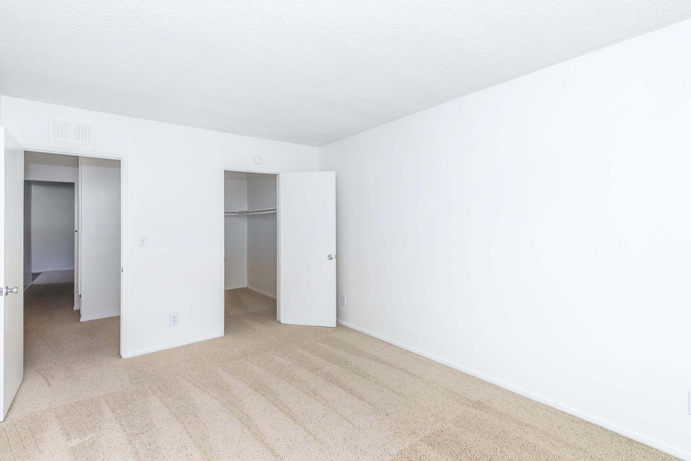 A sparsely furnished room with beige carpet and white walls. One door on the left leads to a closet, while another door on the right opens to a hallway. The room is well-lit and appears empty, providing a blank canvas for potential furnishings and decoration.