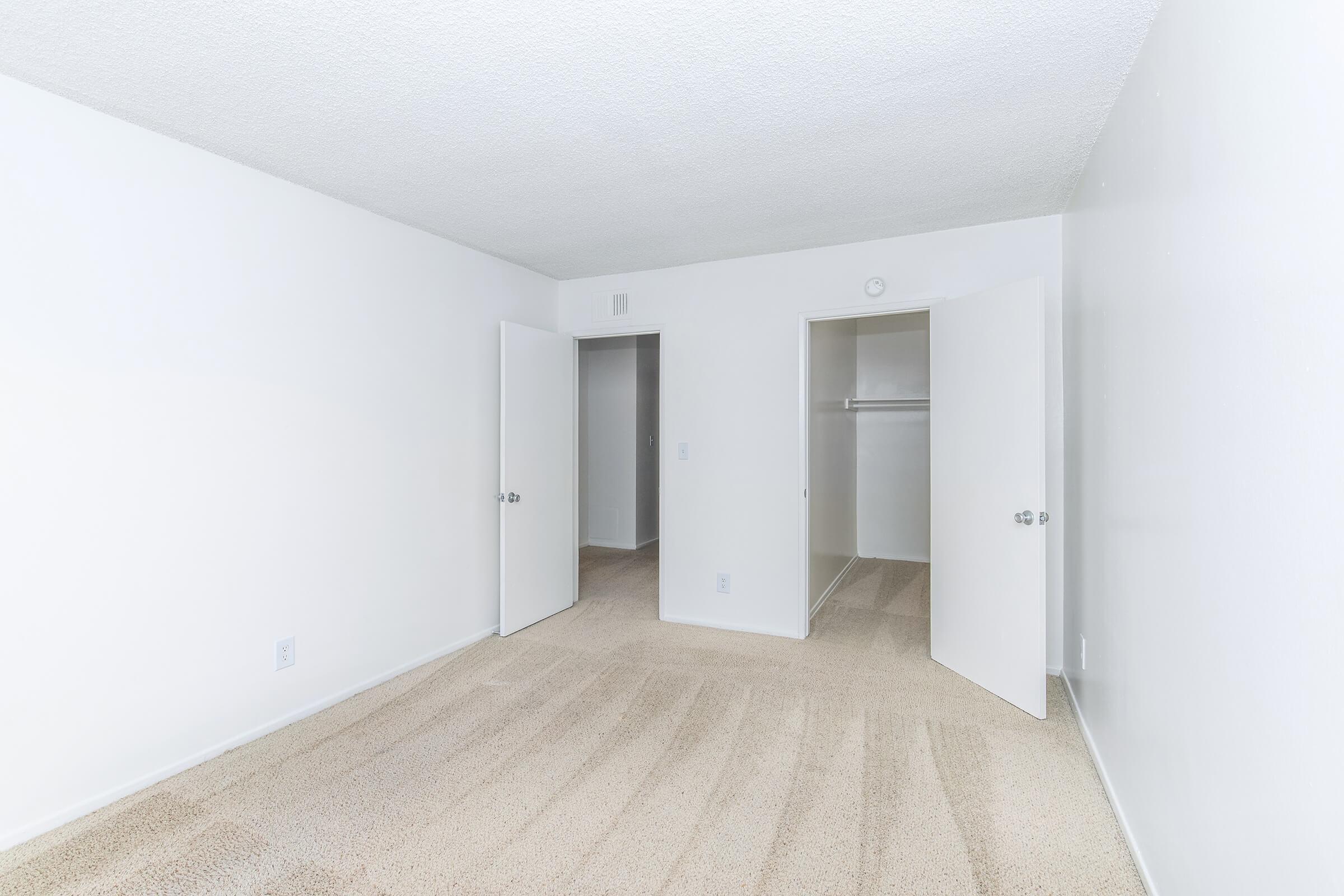 Empty room with light-colored walls and beige carpet. Two doors are open, one leading to a closet with a hanging rod and the other opens to another space. The room has a simple, clean design with ample natural light. No furniture or decorations are present.