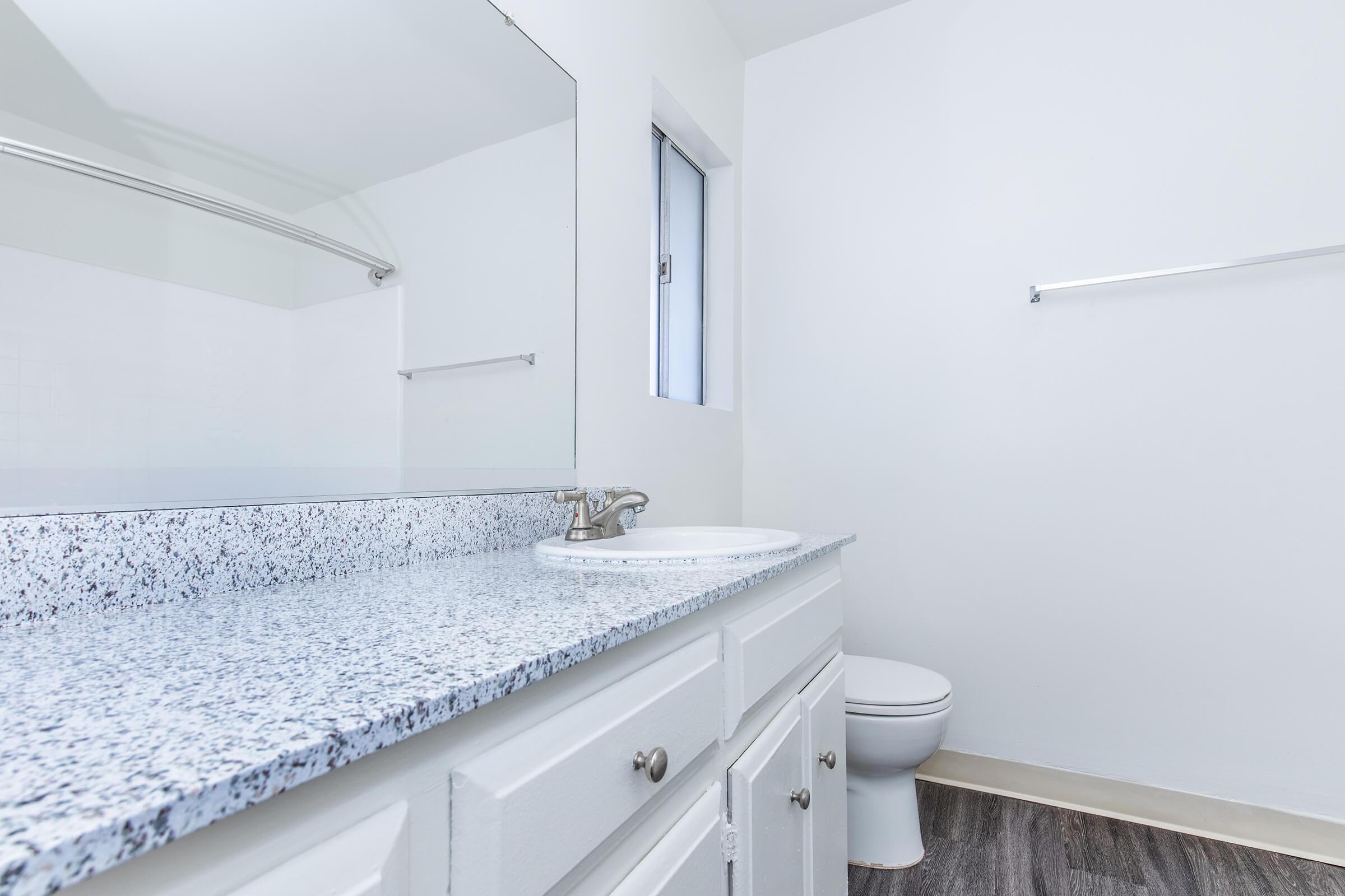 A bright and clean bathroom featuring a white sink with a granite countertop, a mirror above the sink, and a toilet. The walls are painted in a light color, and there is a small window allowing natural light. A towel rack is visible, along with laminate flooring that resembles wood.