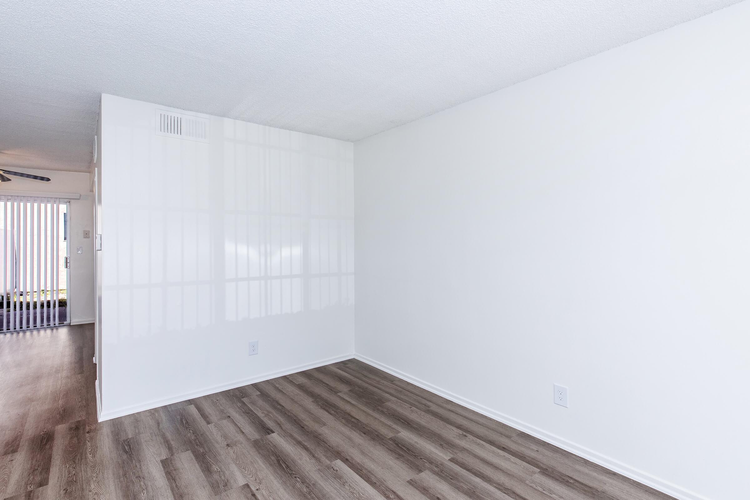 Empty room with white walls and light brown laminate flooring. A window is partially visible on the left side, allowing some natural light. The room has a clean and minimalistic look, with no furniture or decorations. The ceiling is plain and the space feels bright and open.
