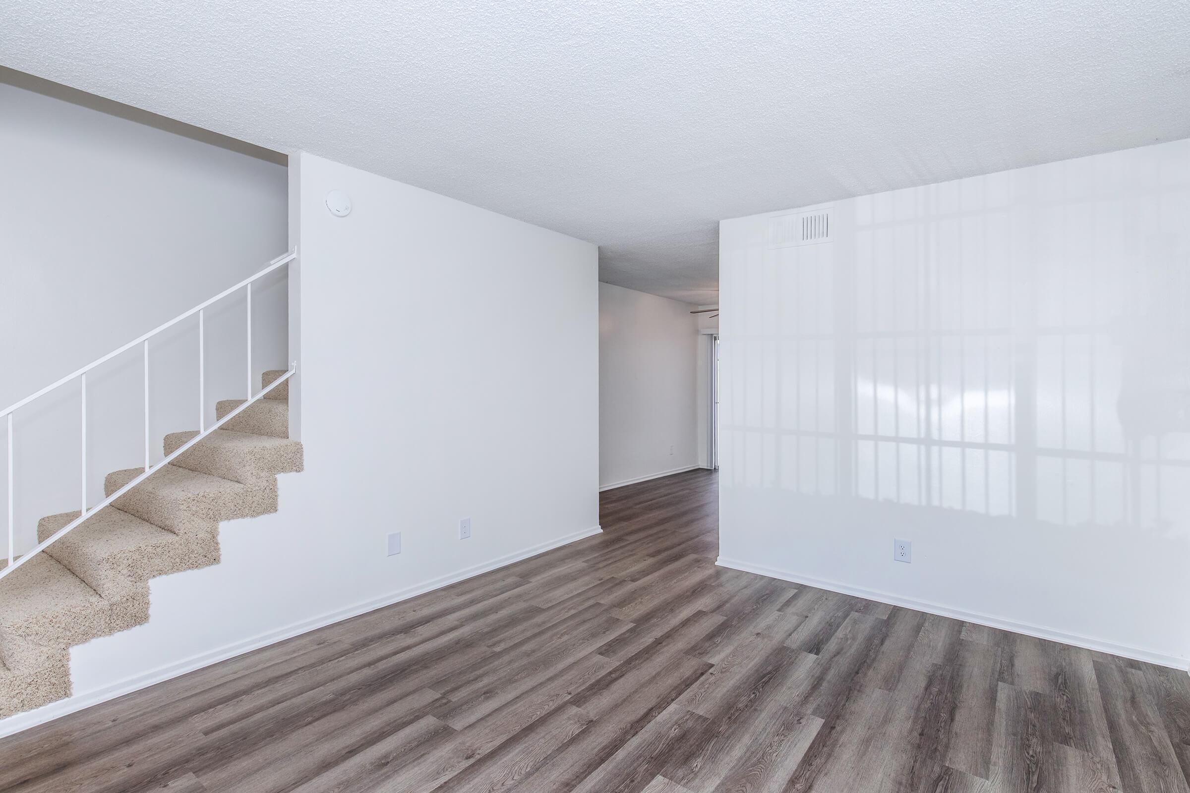 Interior view of a bright, empty living room featuring light-colored walls and wood-like flooring. A staircase with beige carpet is visible on the left side, leading to an upper level. Natural light reflects off the wall, creating a spacious and clean atmosphere.