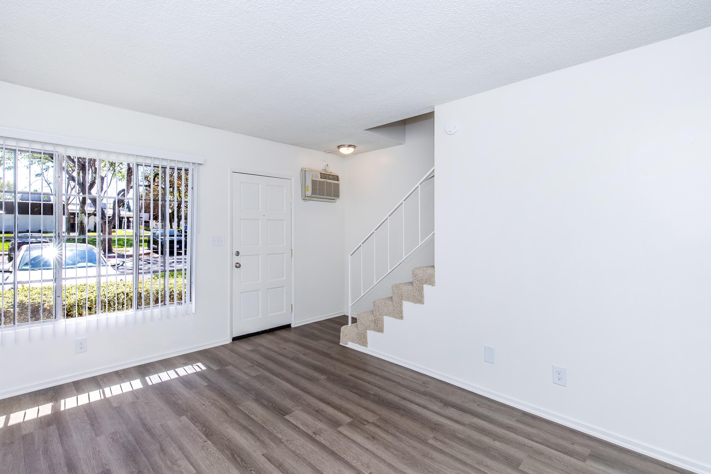 A bright and spacious living room featuring light-colored walls, large windows with blinds, and laminate flooring. A door leads outside while a staircase is visible in the background. The room has minimal furniture, creating an open and airy atmosphere ideal for various decor styles.