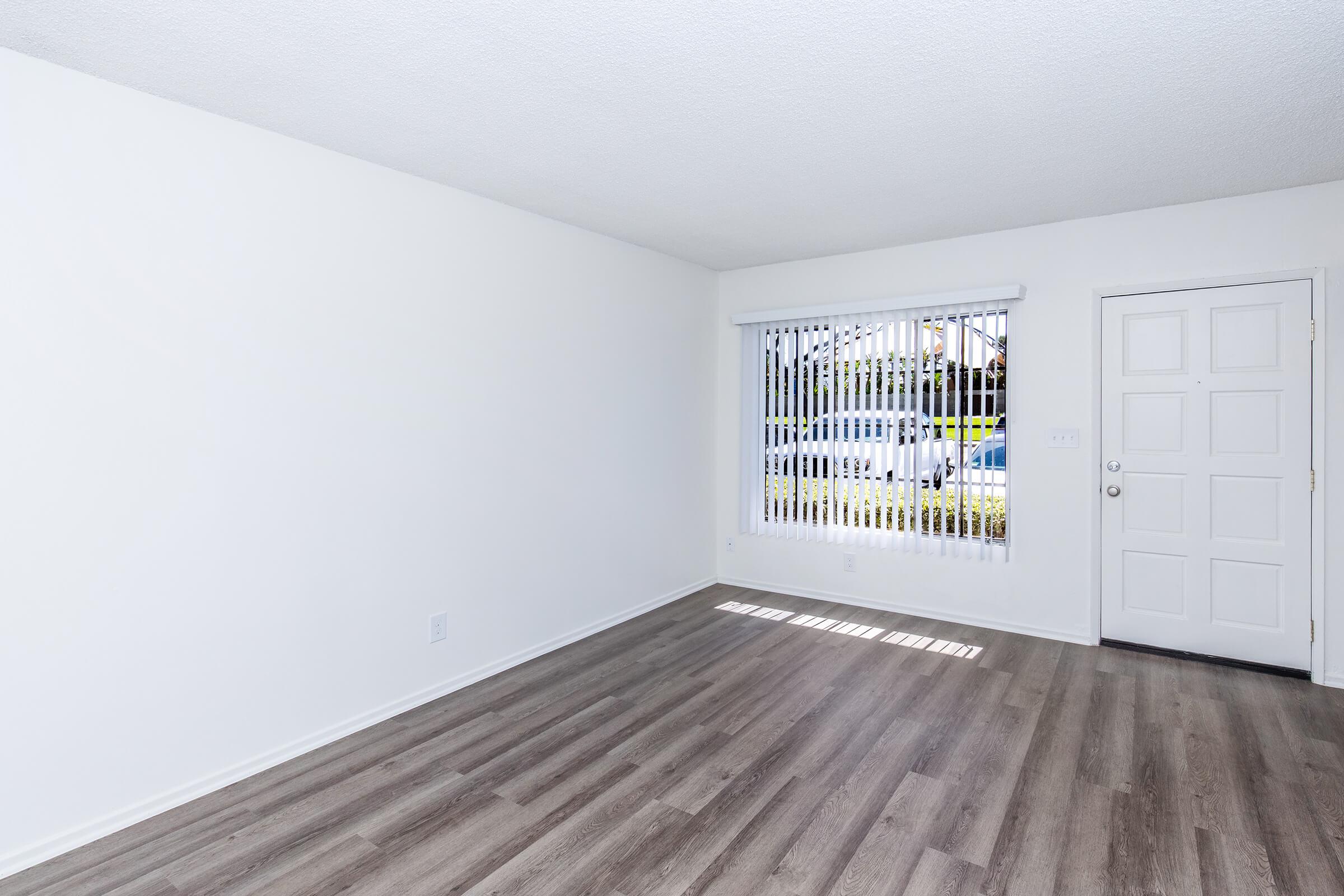 Bright, empty room with light-colored walls and vinyl flooring. A large window with vertical blinds allows natural light to enter, casting shadows on the floor. A white door is visible on the right side of the image, adding to the clean and minimalistic feel of the space.