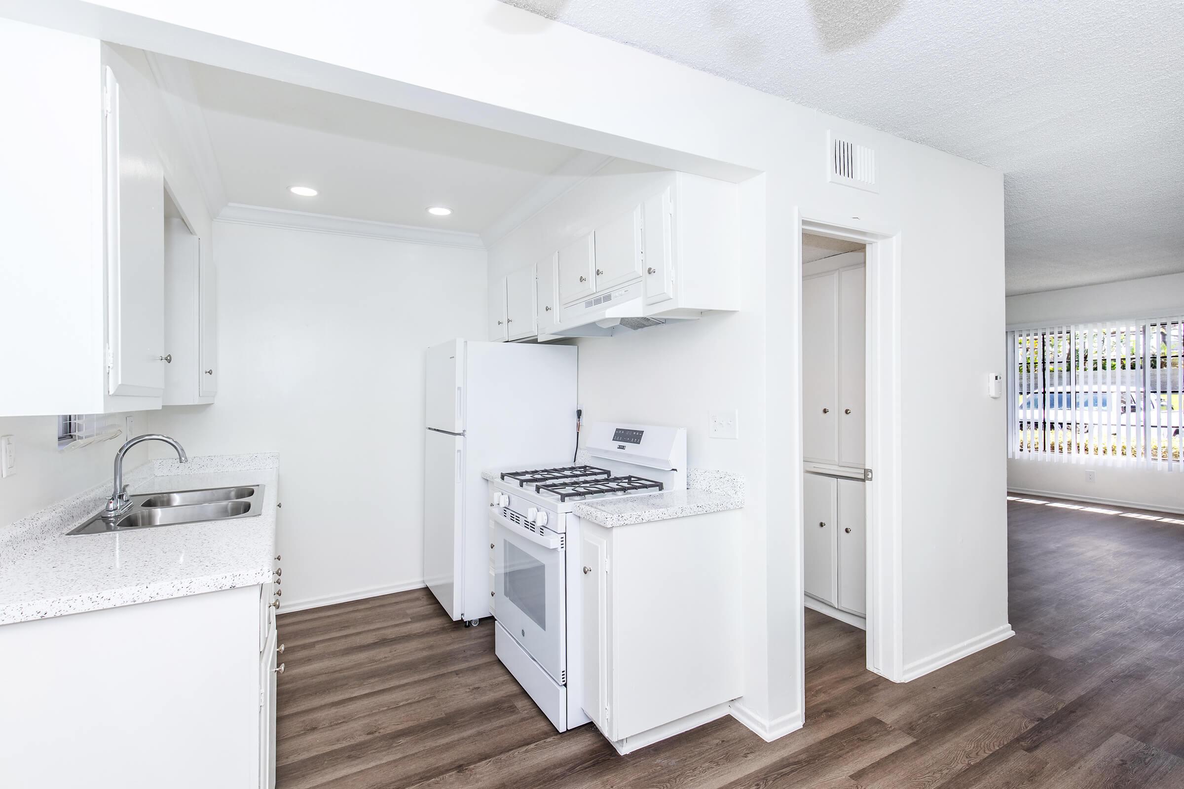 A bright and modern kitchen featuring white cabinetry, a granite countertop, a white gas stove, and a refrigerator. The space has laminate flooring and is well-lit with overhead lights. A doorway leads to another room, with natural light coming through windows in the adjacent area.