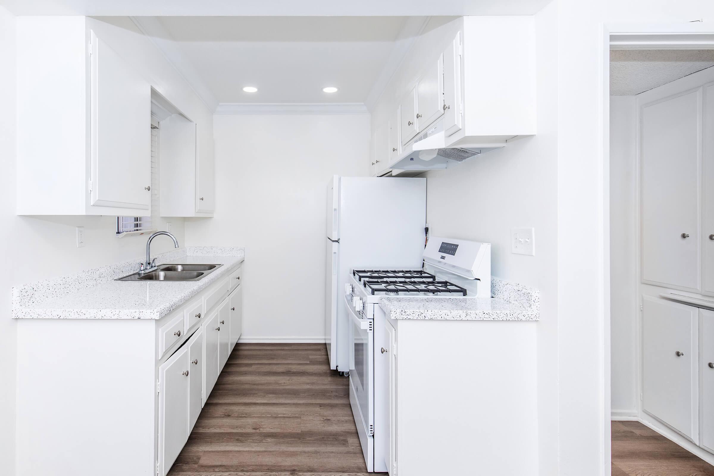 Bright, modern kitchen featuring white cabinets, a stainless steel sink, a white refrigerator, and a gas stove. The countertops are light-colored with a speckled pattern. The space is open and well-lit with natural light coming from windows, showcasing a clean and inviting atmosphere.