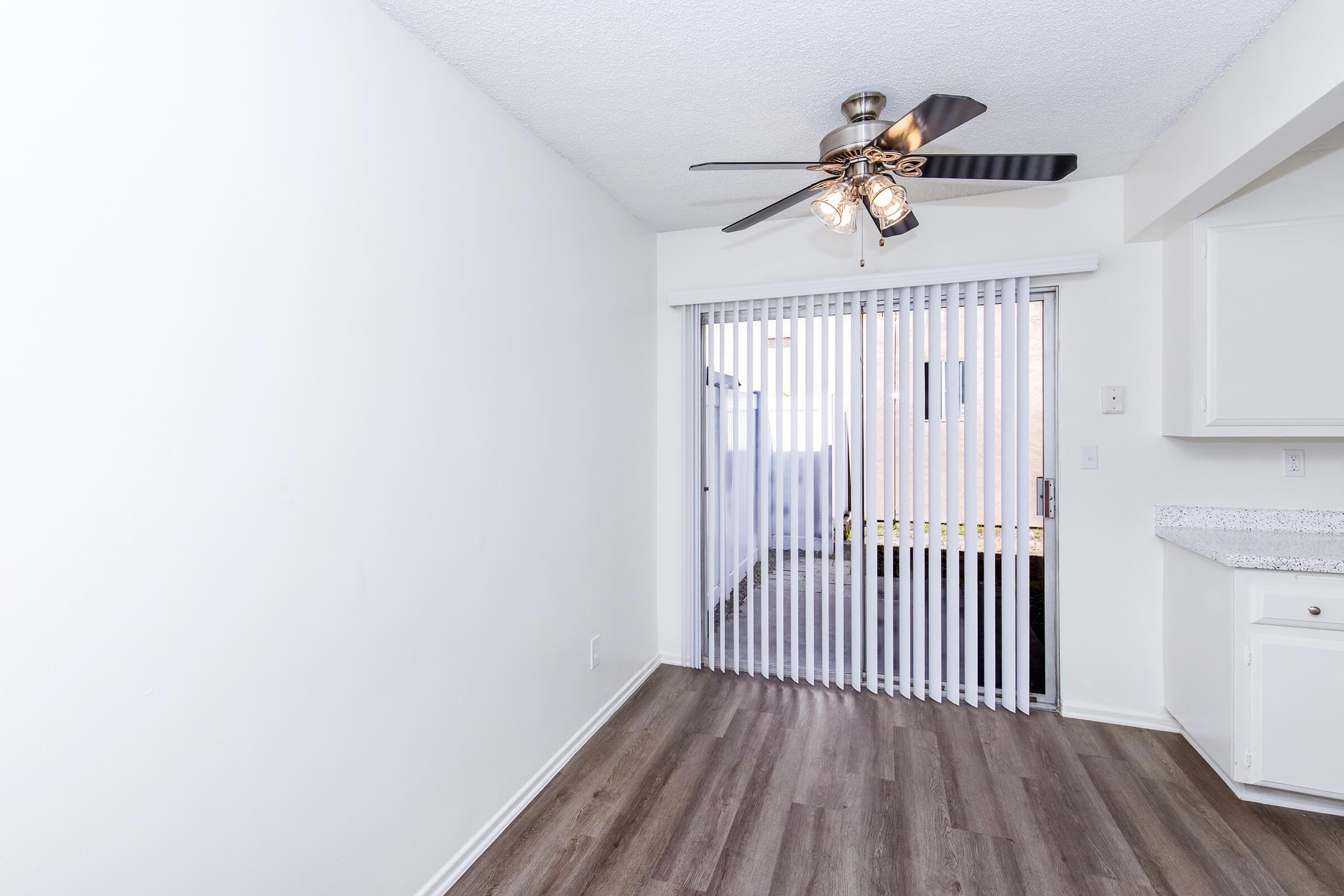 A bright, white room featuring a ceiling fan with lights, hardwood floors, and a sliding glass door covered by vertical blinds that leads to an outdoor area. The walls are plain, and there's a small kitchenette on one side. The space appears clean and modern, ideal for a living or dining area.