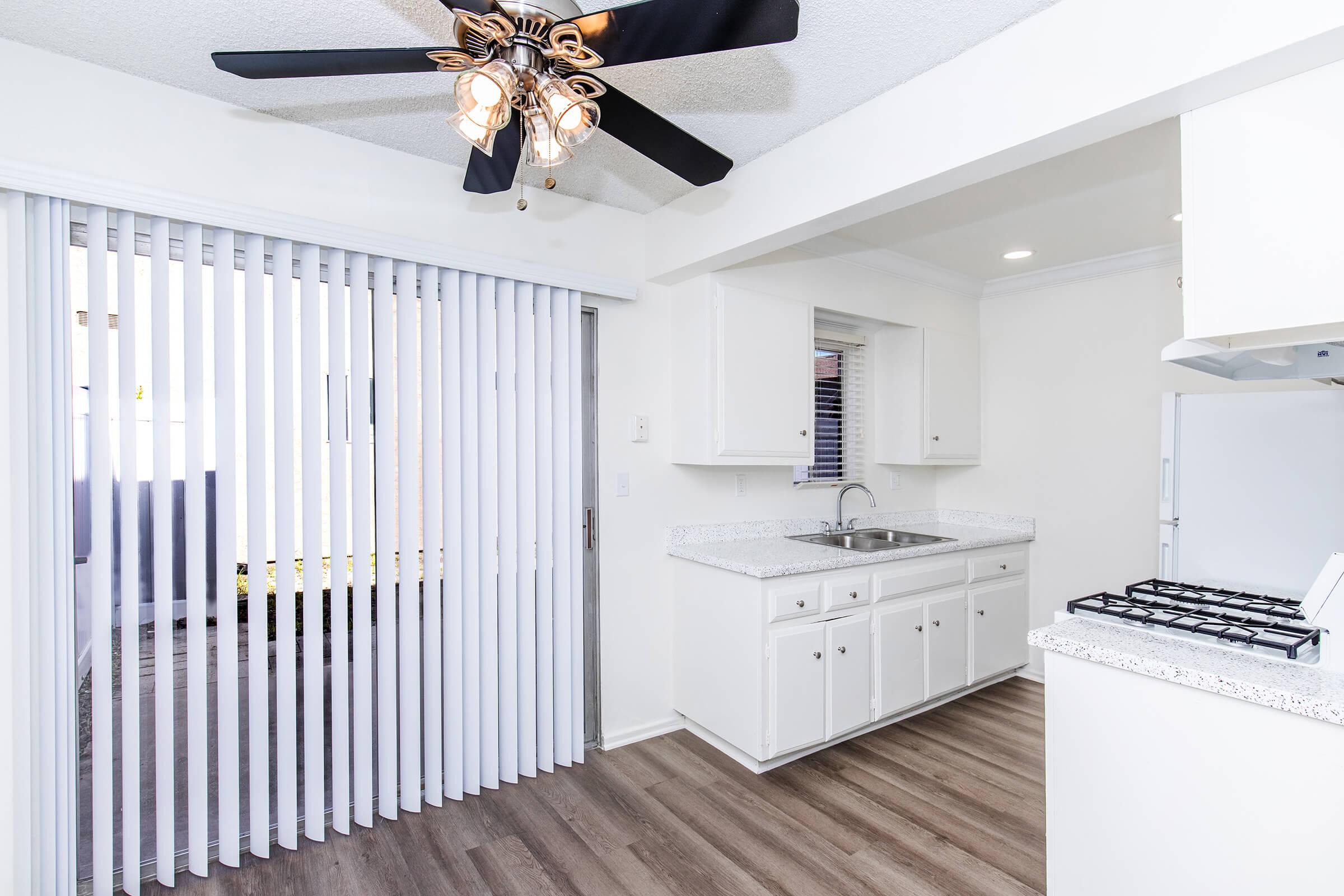 Interior view of a brightly lit kitchen featuring white cabinets, a countertop with a sink, and a gas stove. There's a ceiling fan with lights overhead. A window is visible, along with vertical blinds leading to an outdoor area. The floor is a light hardwood style, contributing to a clean, modern look.