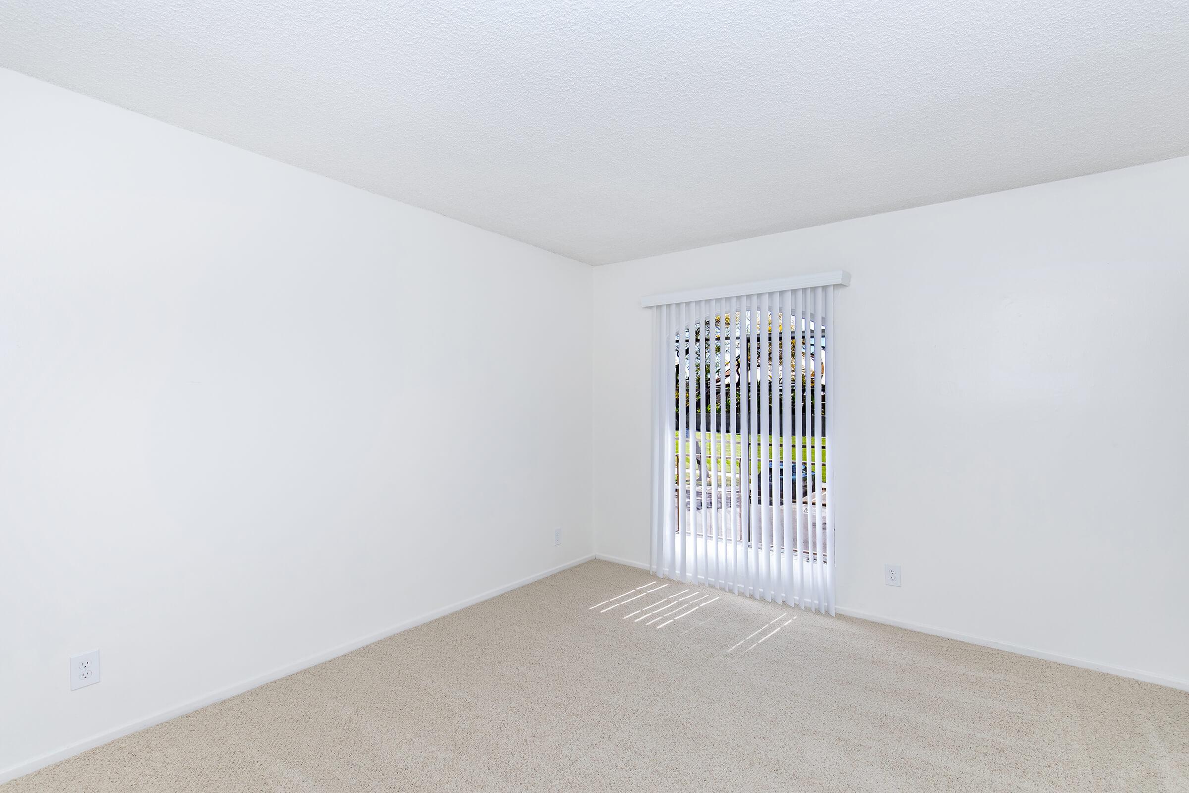 A spacious, empty room with white walls and beige carpet. There's a large window covered with vertical blinds, allowing natural light to enter. Sunlight creates shadows on the floor. The space is clean and uncluttered, ideal for various furniture arrangements.