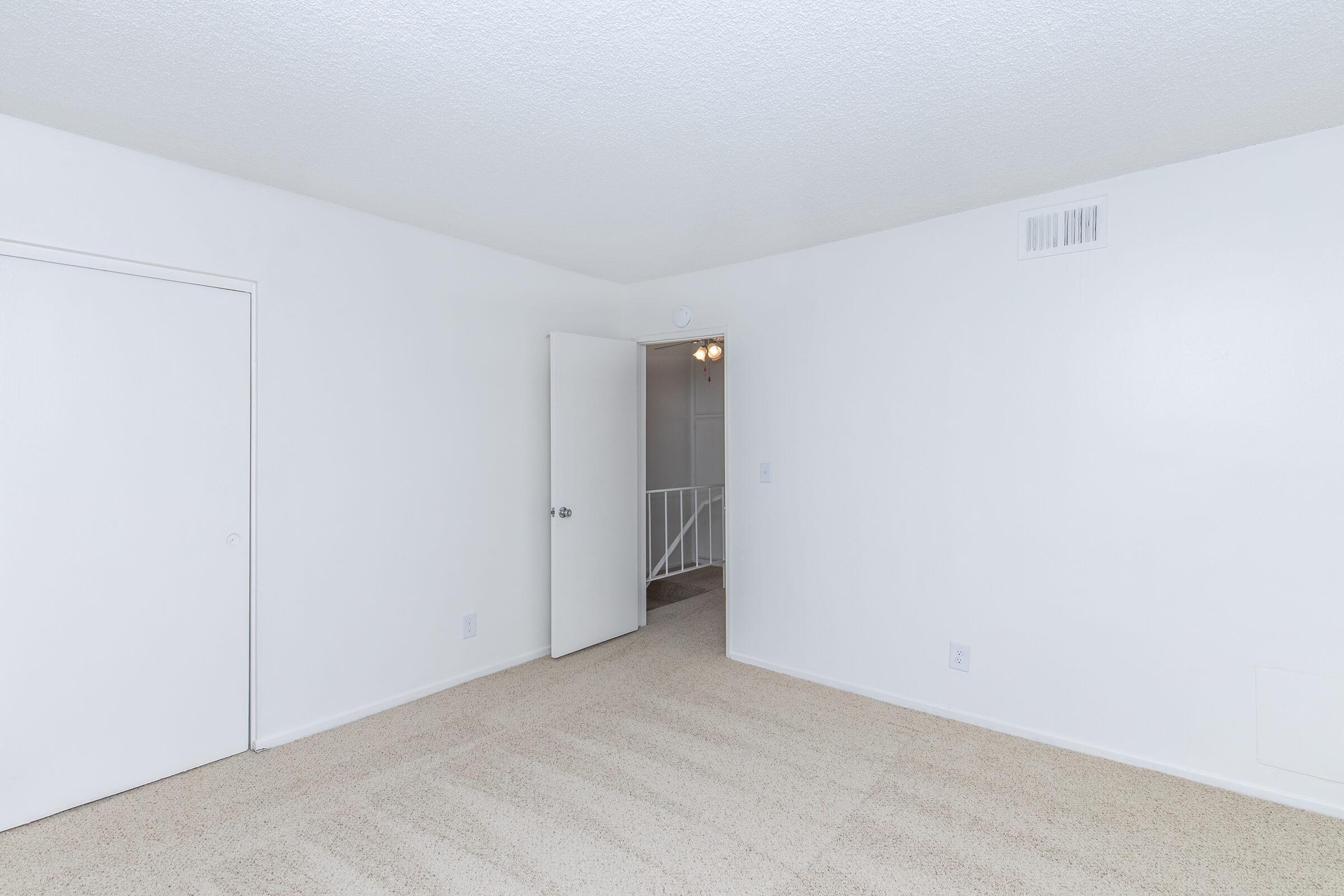 Empty room with white walls and a beige carpet. A closed door is visible to the left, and an open door leads to another area at the back. The ceiling features a light fixture, and there are no furnishings or decorations present, creating a blank canvas environment.