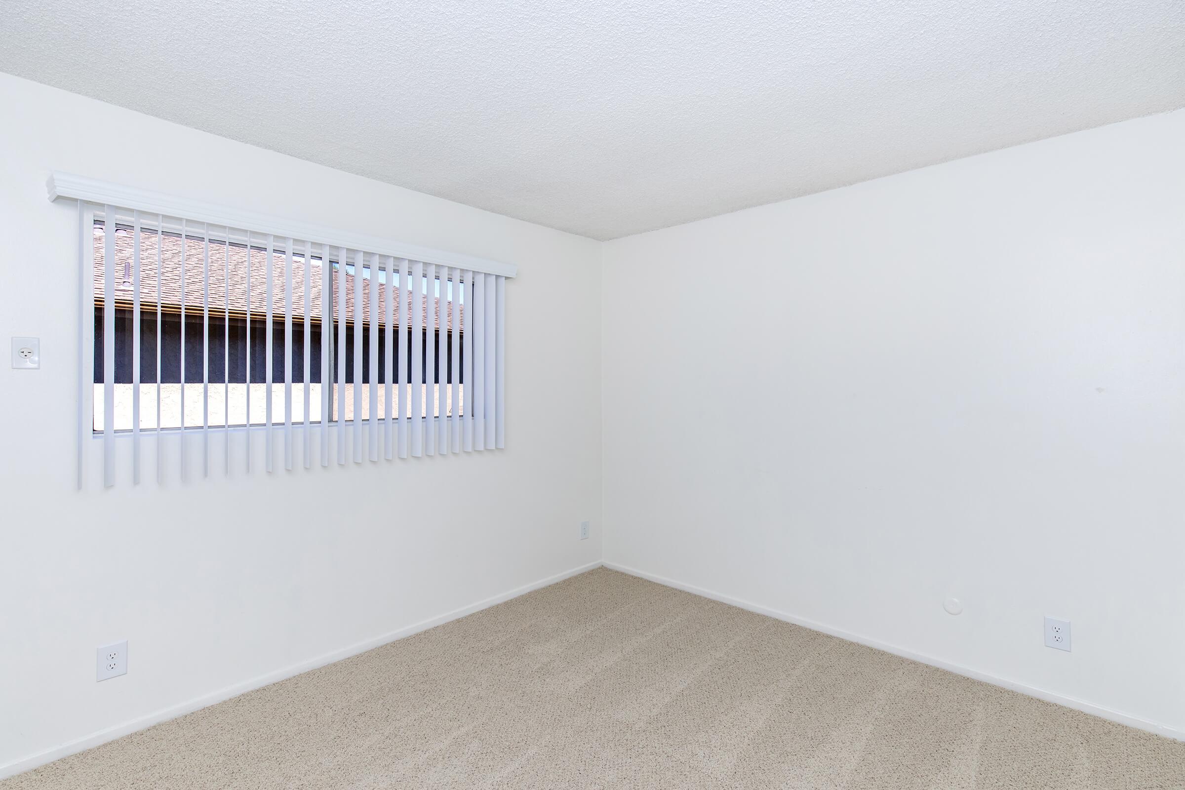 A light-colored empty room with beige carpet and a window covered by white vertical blinds. The walls are painted white, and there is a small electrical outlet on the left side. The window provides natural light and shows a view of an exterior wall outside.