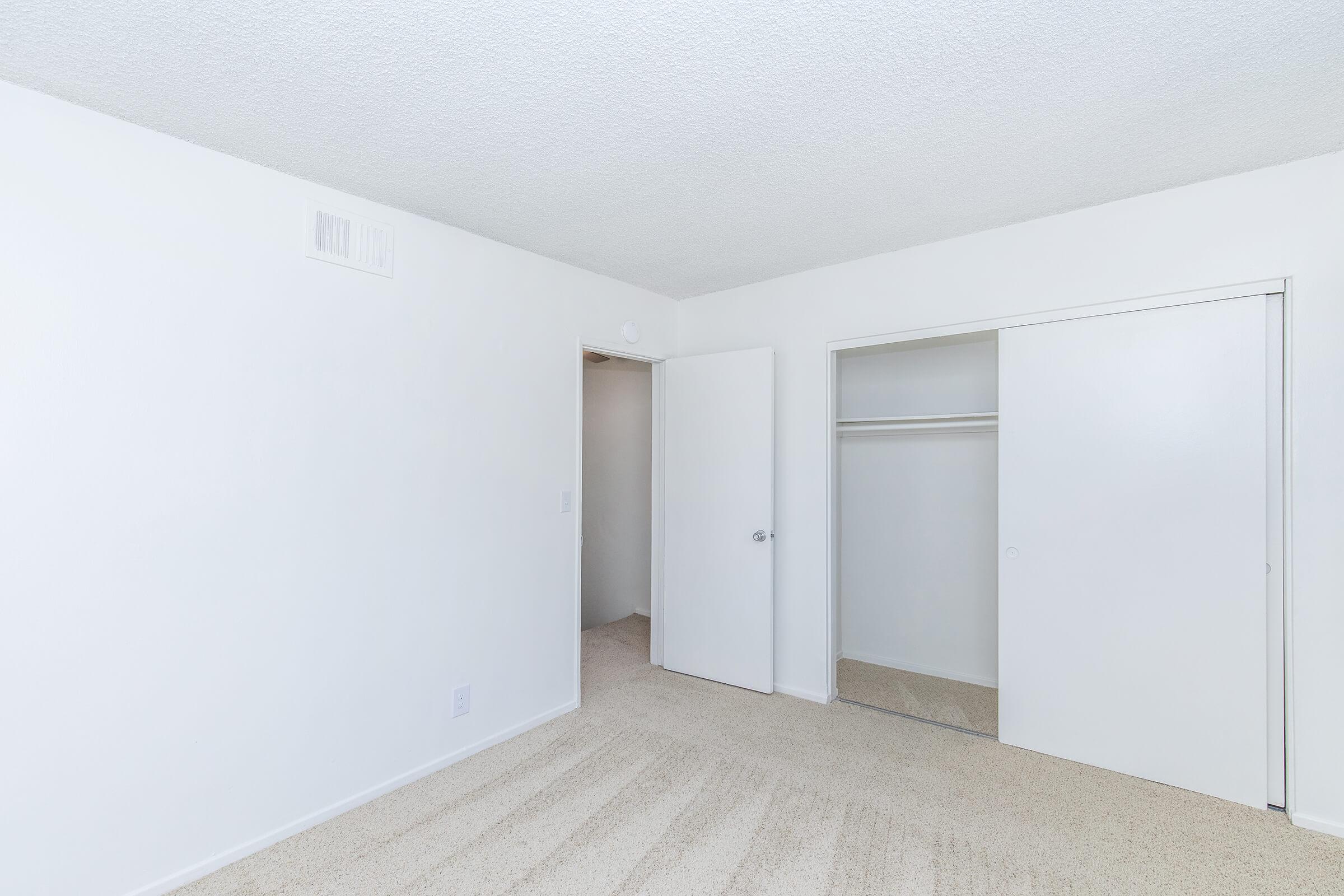 A clean, empty bedroom featuring light-colored walls, carpeted floor, and a sliding closet door. There is a door leading to another room and a small air vent on the wall. The overall atmosphere is bright and minimalist, suitable for personal customization.